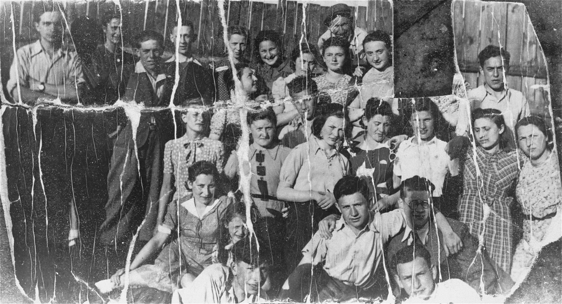 Group portrait of members of the Akiva Zionist youth movement in Tomaszow Mazowiecki. 

The portrait is taken in the yard of Akiva's headquarters on Kryzowa Street.  Pictured from right to left: (top row) unknown, Lonia Lisopraska (cut out), Reginka Orenbach, Motek Kuper (above), Stefa Wygocka, Bronka Plat, Tuska Fuks, unknown, unknown, Halinka Rubinek, Mula Weisman.  Also pictured are Icek Rosenblat (leader of the group), Bronka Rozen, Jadzia Strykowska, Jurek Wald, Chajka Rozenbaum, Aronek Cohen, Lolek Donner.  The donor, Judy Lachman, is the girl directly below the hole. She carried the photo with her throughout the war.