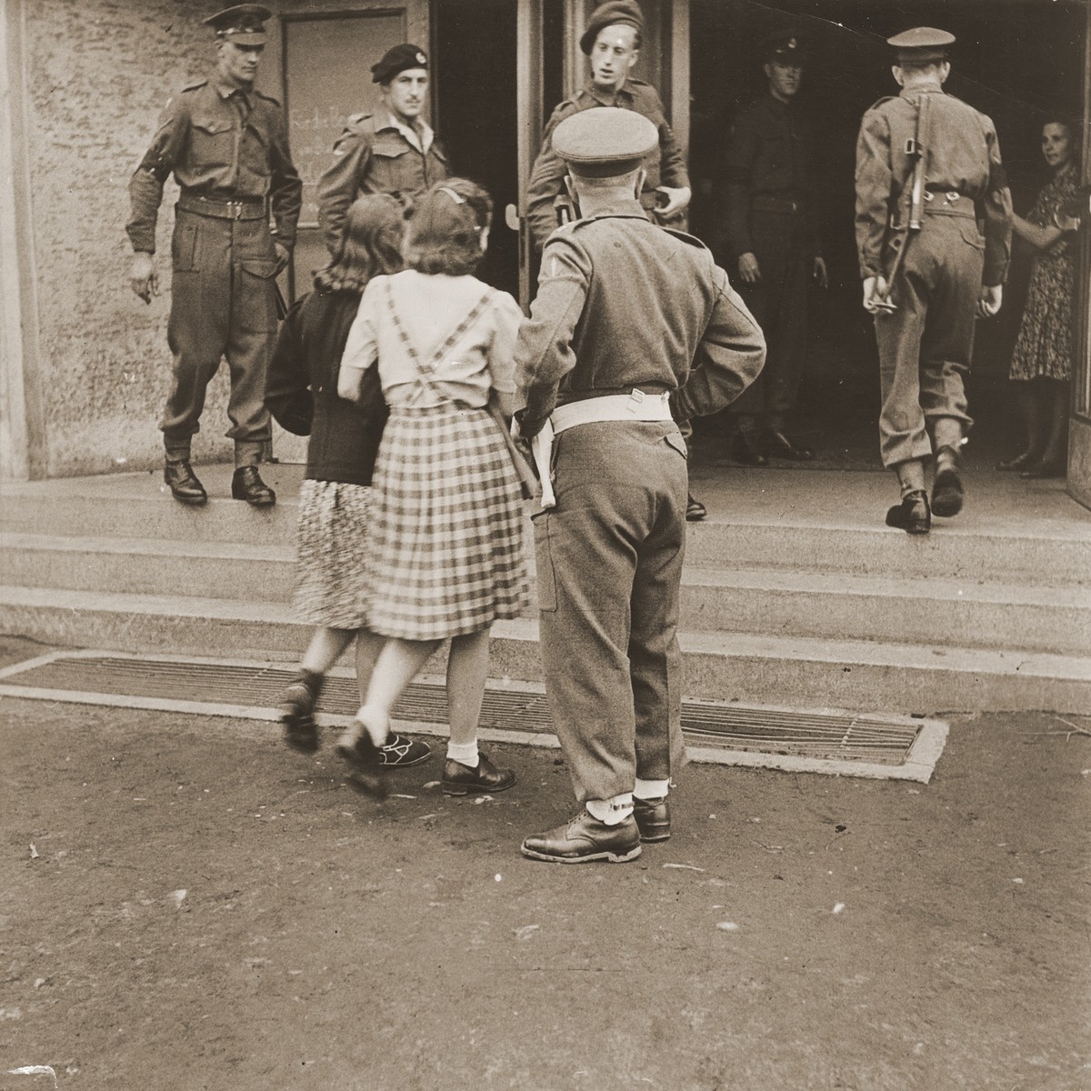 Two German girls from Burgsteinfurt, who laughed when they came out of the cinema showing a film about the concentration camps, are ordered back in to see the film again. 

Burgsteinfurt was called the "village of hate" in the B.L.A. magazine, "The Soldier," because of its silent but noticeable resentment of the British occupation.  The military government began the screening of a documentary on the concentration camps on May 29.  When few residents came voluntarily to view the film, the British military authorities ordered all 4,000 townsmen to attend showings.  They were assembled and marched to the cinema, led by the Mayor and Captain A. Stirling, the District Assistant Provost Marshal.