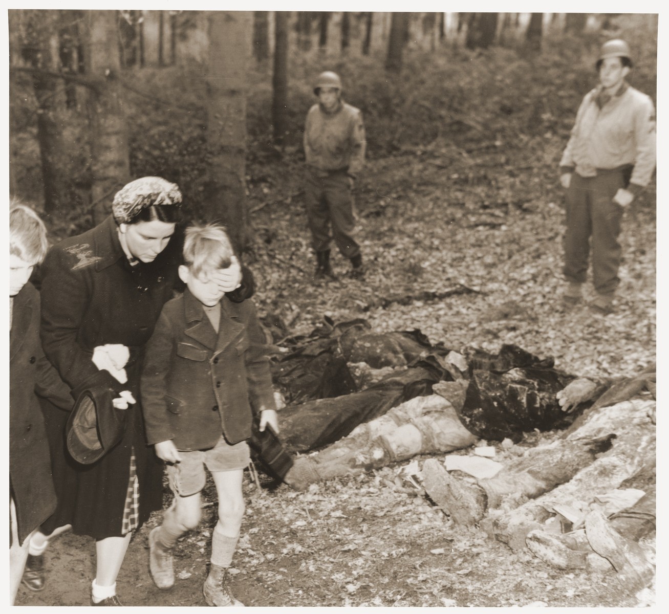 Under the supervision of American soldiers, a German mother shields the eyes of her son as she walks with other civilians past the bodies of 57 Russians, including women and one baby, exhumed from a mass grave outside the town of Suttrop.  

The victims were forced to dig their own grave and then were shot by SS troops six weeks before the arrival of American troops.  On May 3, 1945, the 95th Infantry Division of the U.S. Ninth Army arrived in Suttrop and were informed by locals of the mass grave.  American troops forced the townspeople to exhume the grave after which Russian displaced persons in the area identified the bodies.  The victims were reburied in individual graves, and a U.S. Army chaplain conducted burial services.  Russians remaining in the area placed wreaths on the graves.