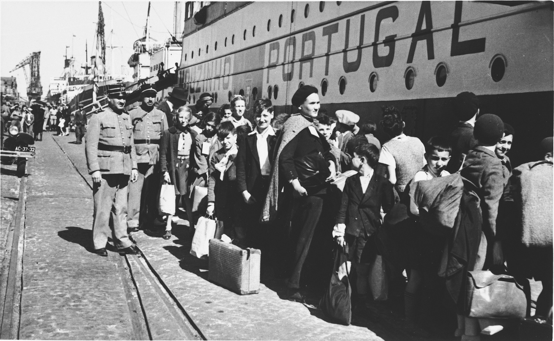 Two uniformed Portuguese policemen stand on the pier in the port of Lisbon as a group of Jewish refugee children wait in line to board the SS Mouzinho.

The young boy just to the right of center in the image, looking back over his right shoulder, is Herman Rosenfeld, born 27 April 1933 in Adelheim, Germany. 
The young blonde girl standing next to the two police officers is wearing tag #25, can be identified as Lilian Warschawski (or Warszawski), born 20 April 1930 in Belgium.