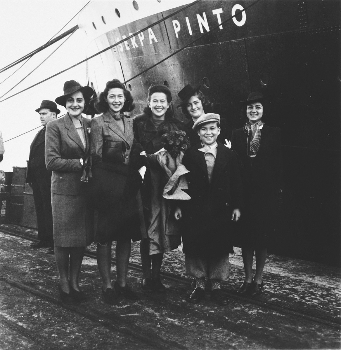 A group of Jewish refugees pose on the pier in the port of Lisbon before boarding the SS Serpa Pinto. 

Among those pictured is Madeleine Hecht Feher, center holding a bouquet of flowers, surrounded by her co-workers from the American Joint Distribtuion Committee.  They came to wish her farewell before embarking.  Also pictured is her brother, Thomas Hecht, the young boy in front.   Pictured on the far left is Lolita Goldstein, a JDC worker.
