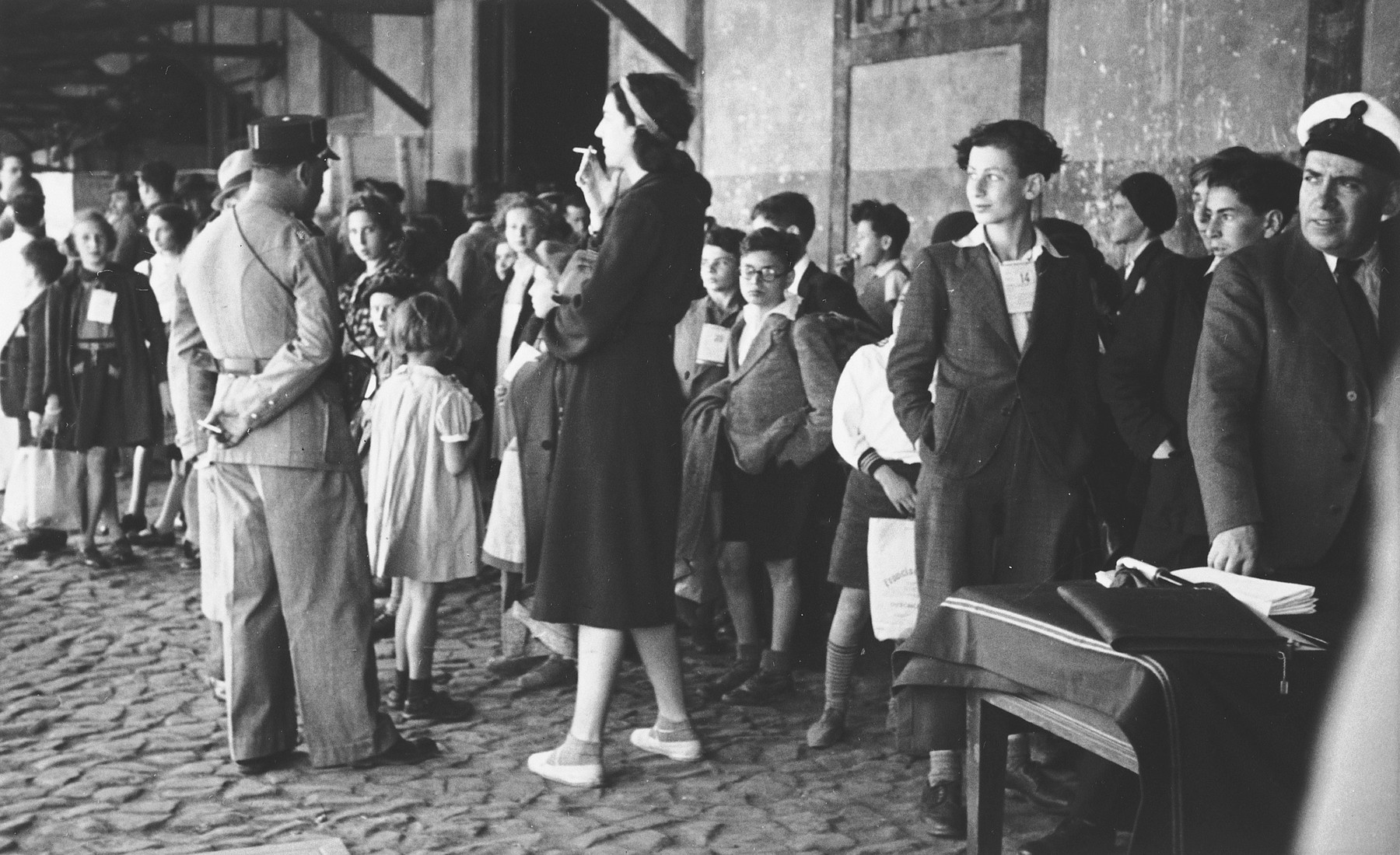 A group of Jewish refugee children wait in the port of Lisbon to board the SS Mouzinho.

On the right side of the photo a young man is wearing a tag clearly legible as #14. This is Guenther Sinasohn born 29 October 1926 in Germany. There was also a Herbert Schwartz (born 16 May 1925) on this transport who may have been Guenther's brother.The young blonde girl on the far left of the image is wearing tag #25, and can be identified as Lilian Warschawski (or Warszawski), born 20 April 1930 in Belgium.  In the middle of the photograph the young boy wearing what appears to be ID tag #23 is Oswald Kernberg, born 19 October 1929 in Vienna.  Pictured in the back, fourth from the right in profile is  Alexander Ssascha Britan. The tall woman smoking a cigarette might be Ursula Gajewska, who was the adult escort on this transport. Ursula was an American citizen, born 1908 in Buffalo, NY, who was fleeing Poland herself.