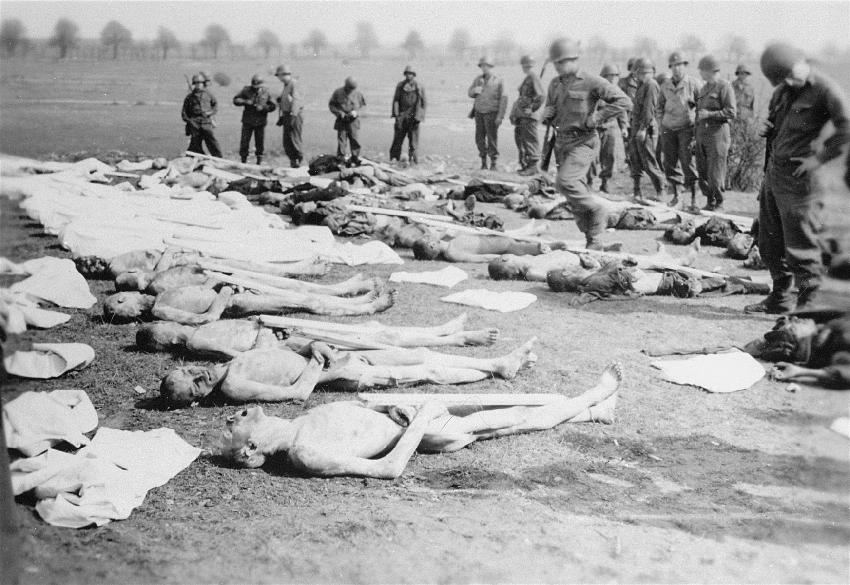 American soldiers stand among corpses laid out for burial by German civilians conscripted to clean up the Ohrdruf concentration camp.  Due to a lack of information about the names of the dead, each body was assigned a number to be placed on the grave marker.