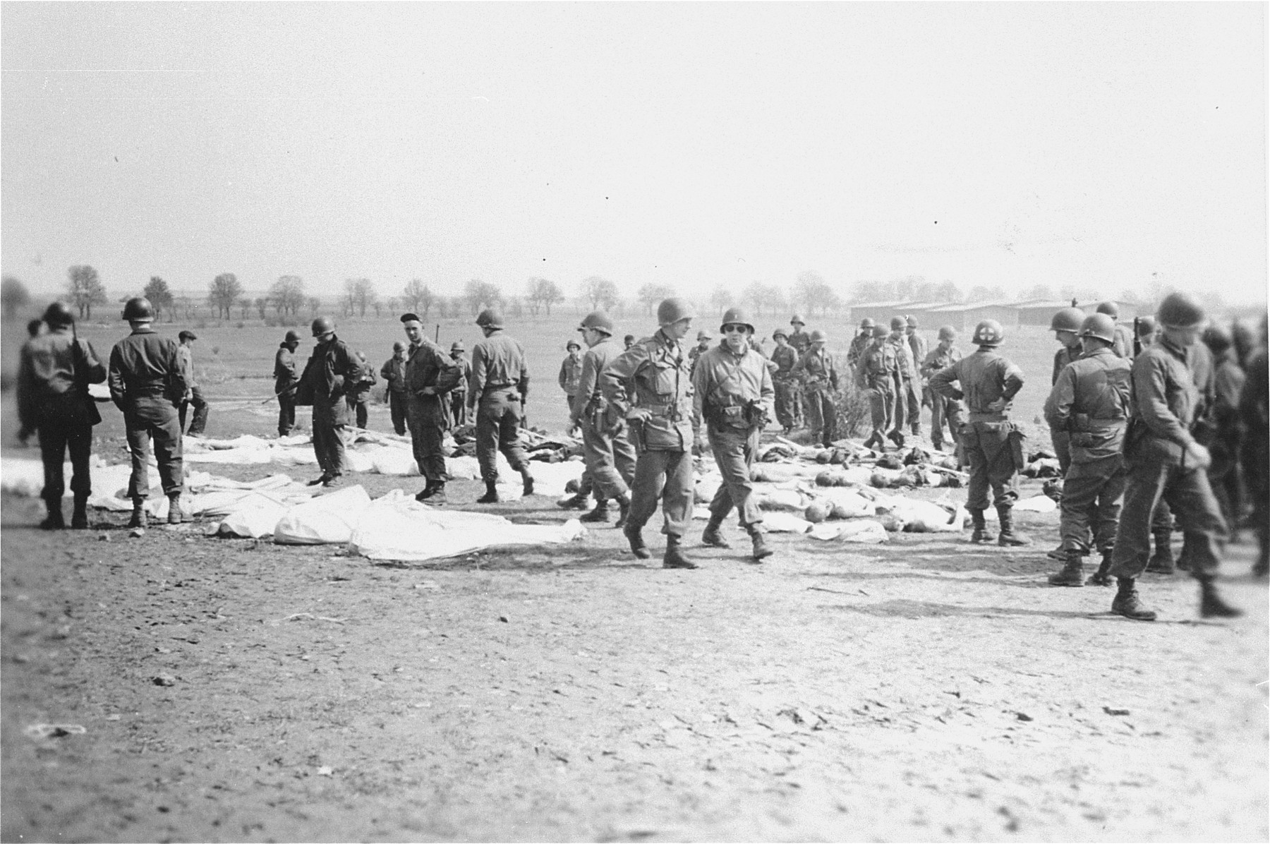 American soldiers stand among corpses laid out for burial by German civilians conscripted to clean up the Ohrdruf concentration camp.  Due to a lack of information about the names of the dead, each body was assigned a number to be placed on the grave marker.