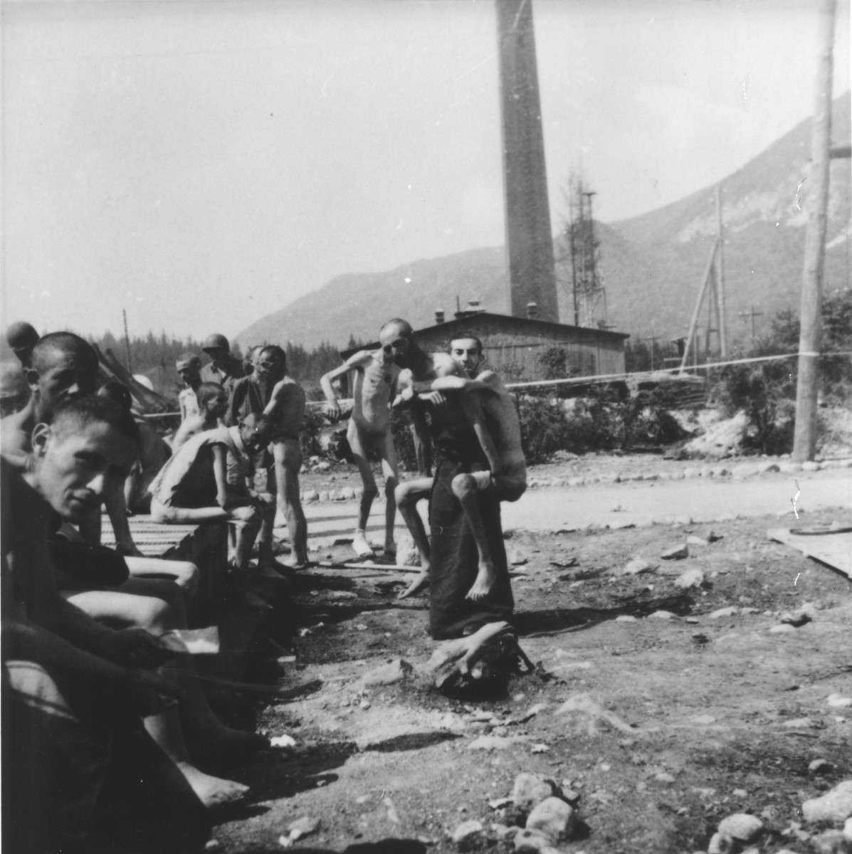 A survivor carries a weaker fellow to the latrine, which American soldiers built for them.  The camp crematorium looms in the background.