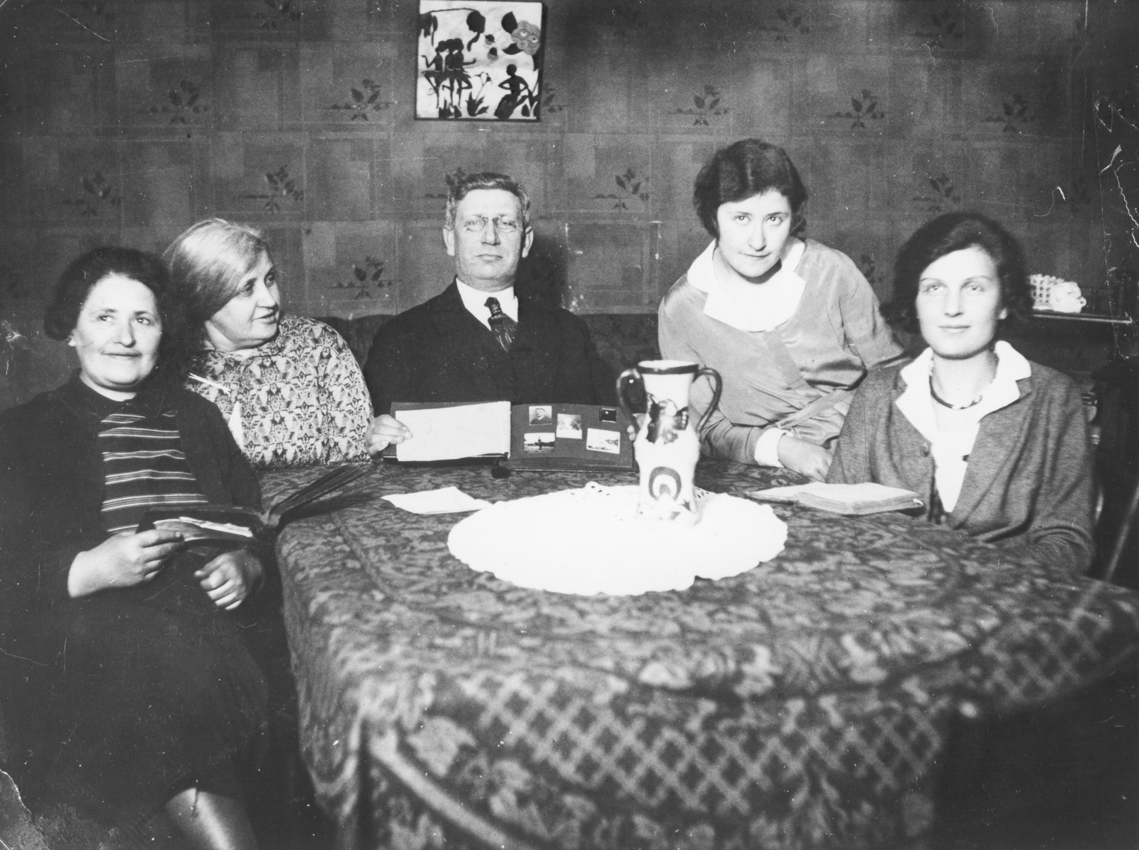 Members of the Magid family sit around their dining room table, displaying a photo album.

Pictured are Genia and Boris Magid (second and third from the left), Mania Shambadahl Olitsky and Katia Magid (right).