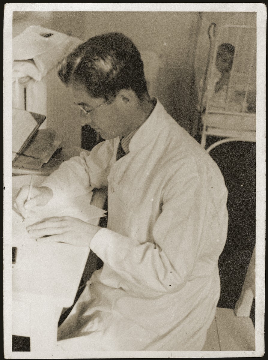 A Jewish pediatician writes at his desk while a young child sits in a crib behind him.
