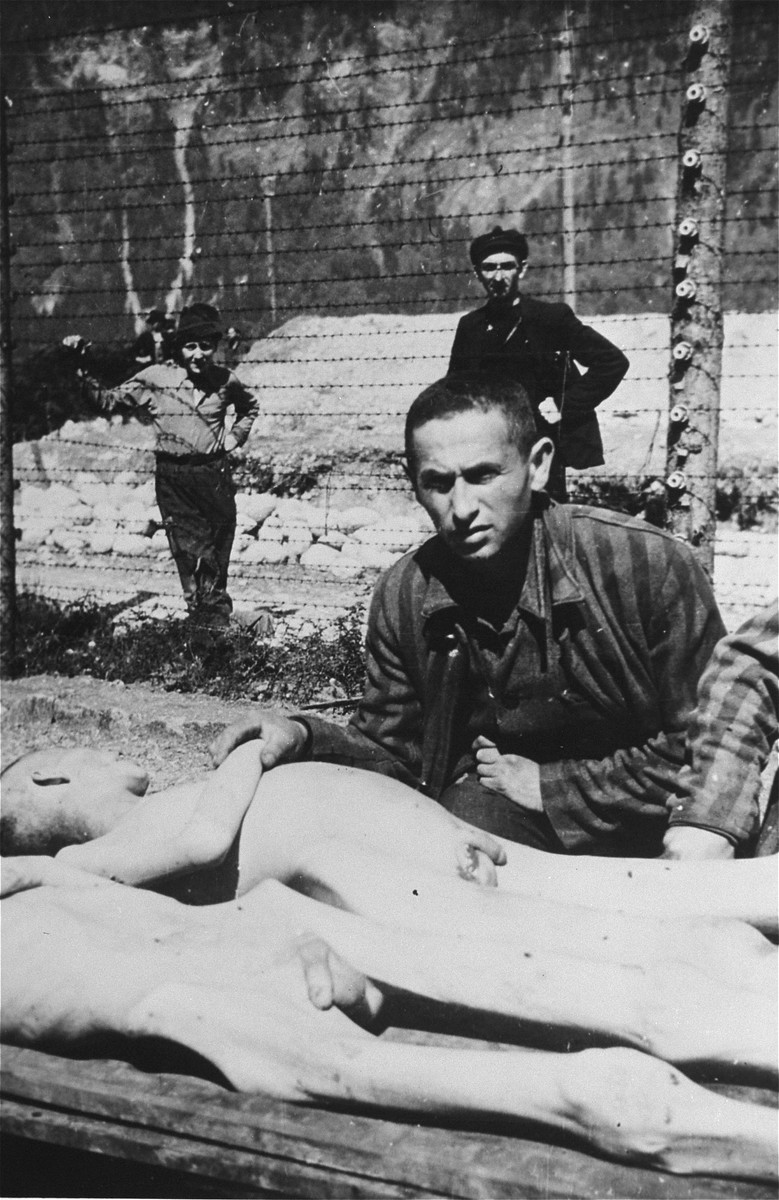 A survivor leans over the corpses of two fallen comrades after liberation. Two survivors look on through the barbed wire fence.