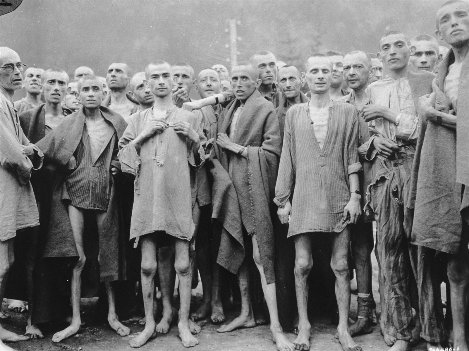 One day after their liberation, a group of former prisoners at the Ebensee concentration camp pose outside for US Army Signal Corps photographer Arnold Samuelson.

The former prisoners came out of the infirmary barracks in order to be photographed.  The man in the center left holding his metal name tag has been identified as 21-year old Jewish survivor Joachim Friedner from Krakow, Poland.  The man in the center back, holding up his arm to show his arm band, has been identified both as William Hauben and as Isachar Herszenhorn (later Irving Horn) from Radom, Poland. Bernard Smilovic, from Munkacs, is located second from the left, first row wearing a blanket.  The man in the front, center , wrapped in a blanket, has been identifided as Gustav Fabik. He was born on September 5th 1917. He was captured on November 4th 1944 and sent to Dachau. He was transported to Mauthausen and liberated on May 5th 1945.  In the back row, on the far right, is Zev Harel.  

Original caption reads: " Starved prisoners, nearly dead from hunger, pose in the Nazi concentration camp in Ebensee, Austria.   In the Austrian Alps the Nazis had one of their largest camps.  Large numbers of inmates were starving to death and dying at the rate of 2000 per week.  The camp was reputedly used for "Scientific" experiments.  It was liberated by the 80th Division, 3rd U.S. Army."


Original caption from donated photograph reads:  "Starved prisoners, nearly dead from hunger, pose for the photographer in their rags."