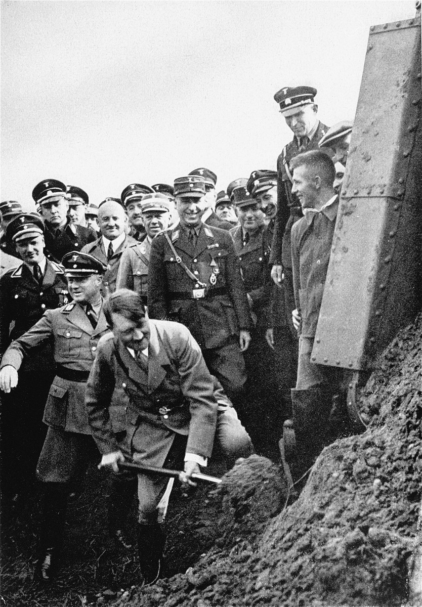 Adolf Hitler inaugurates the construction of a stretch of the Autobahn by turning over the first shovelful of earth, as Nazi officials look on.