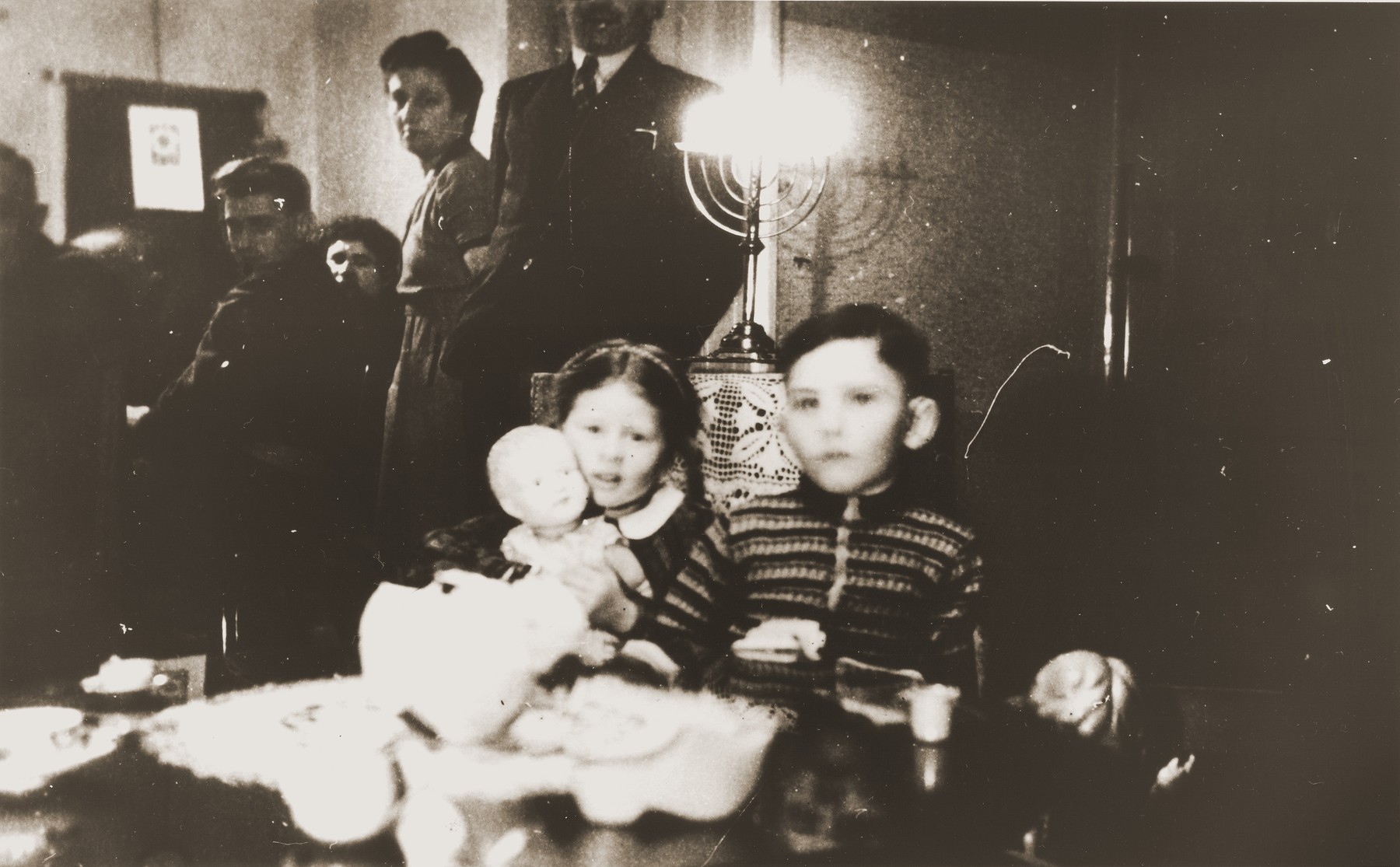 Two young Jewish children sit in front of a lighted Hanukkah menorah at the first postwar family Hanukkah celebration at the Zion home in Eibergen.

Pictured clutching her new doll is Betty Rosenbaum.  Betty, who was orphaned during the war, was adopted by her uncle Julius Zion after his marriage in 1946.