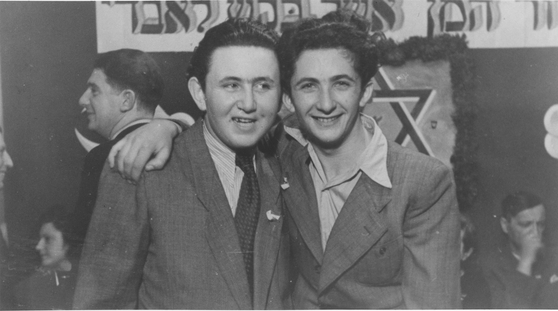 Two young men pose at a Purim party in the Schlachtensee displaced persons camp.

Yeshayahu Zycer (left) and Mischa Gleiberman (right) stand beneath a Hebrew banner that reads, "Cursed be Haman who tried to kill me." (a quotation from the Book of Esther recited on the holiday of Purim).