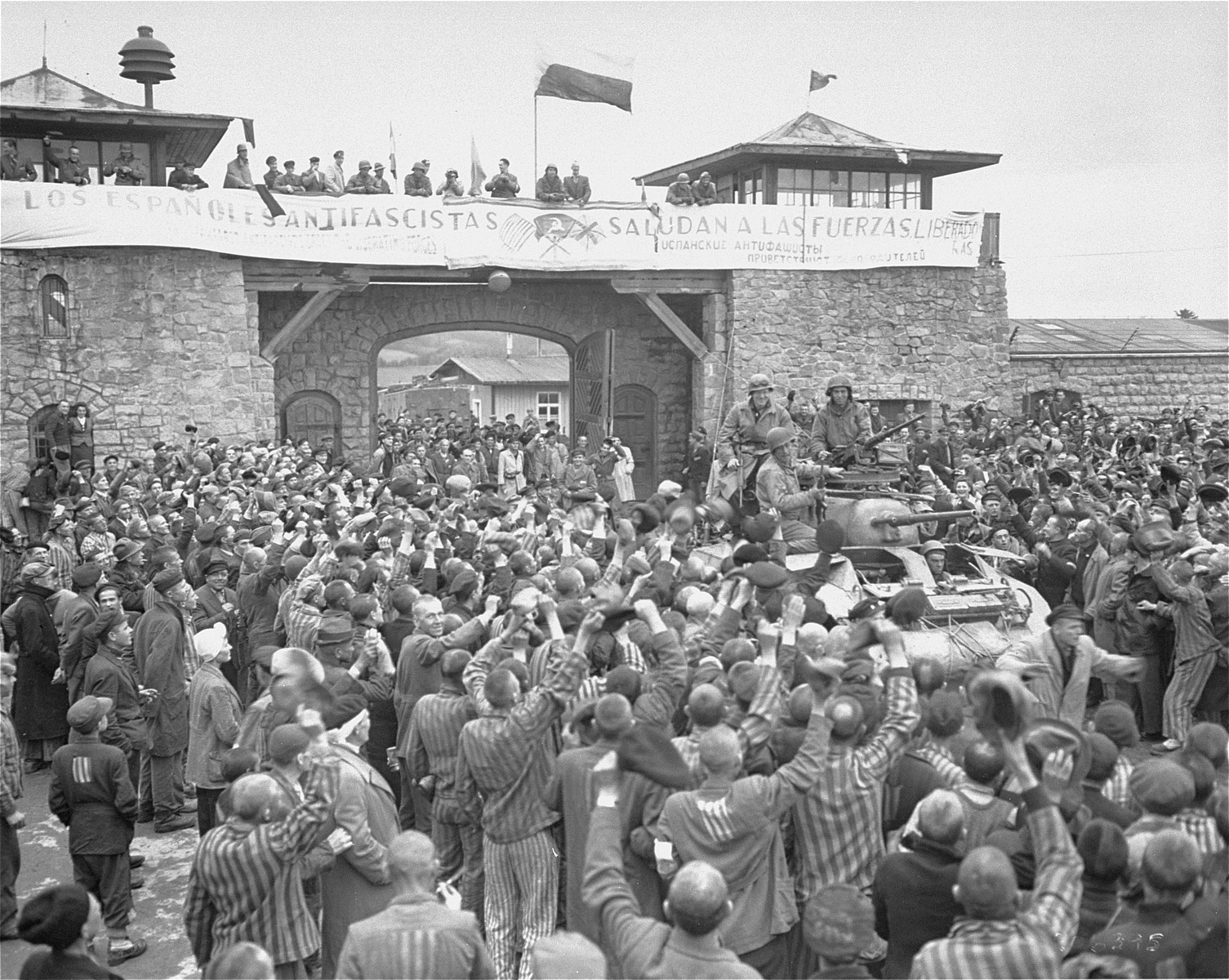 Mauthausen survivors cheer the soldiers of the Eleventh Armored Division of the U.S. Third Army one day after their actual liberation. 

The banner reads: "The Spanish Anti-Fascists Salute the Liberating Forces."  According to Pierre Serge Choumoff, a survivor of Mauthausen and Gusen, this event was recreated at the request of a senior American officer.

The photographer Francisco Boix can be seen with his camera standing on top of the entrance.  According to the 11th Armored Division website the solders are John Slatton (back left), Jerome Rosenthal (back right), William Picket (left) and Edward Czarnowski (driver).  Alternatively Robert Mordis, a Jewish soldier from Natick Massachusetts, may be on the upper left side of the tank, and another soldier may be Alfred Paliani.