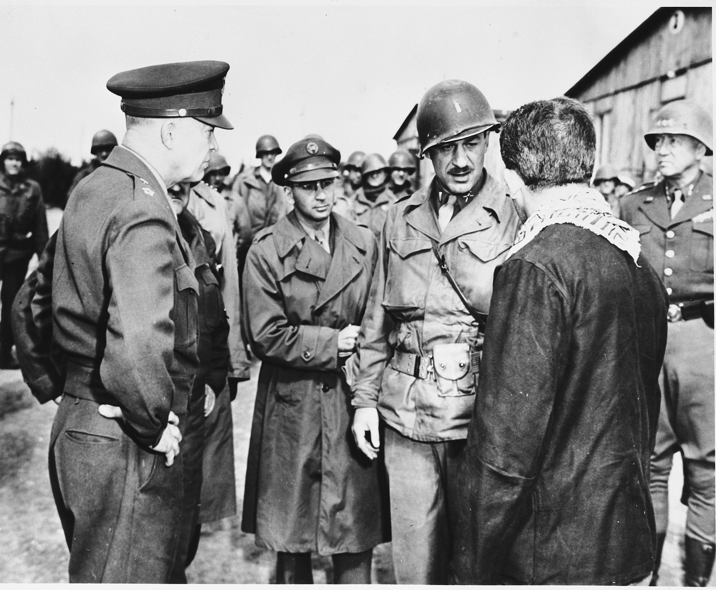 An Austrian-Jewish survivor talks with high-ranking U.S. Army officers in the newly liberated Ohrdruf concentration camp.

Among those pictured are General Dwight D. Eisenhower (left), and General George Patton (right).  Also pictured is Jules Grad, correspondent for the U.S. Army newspaper, "Stars and Stripes" (center).  The Austrian Jewish survivor is Ignaz Feldmann (wearing a dark coat, with a scarf around his neck) . He was a survivor of  Westerbork, Terezin, Auschwitz, and Ohrdruf camps.   He was also a player on the famed Hakoah Vienna soccer teams of the 1920s and 30s.

The mustached soldier is Alois J. Liethen of Appleton, WI, who served as the interpreter for the tour of Ohrdruf.