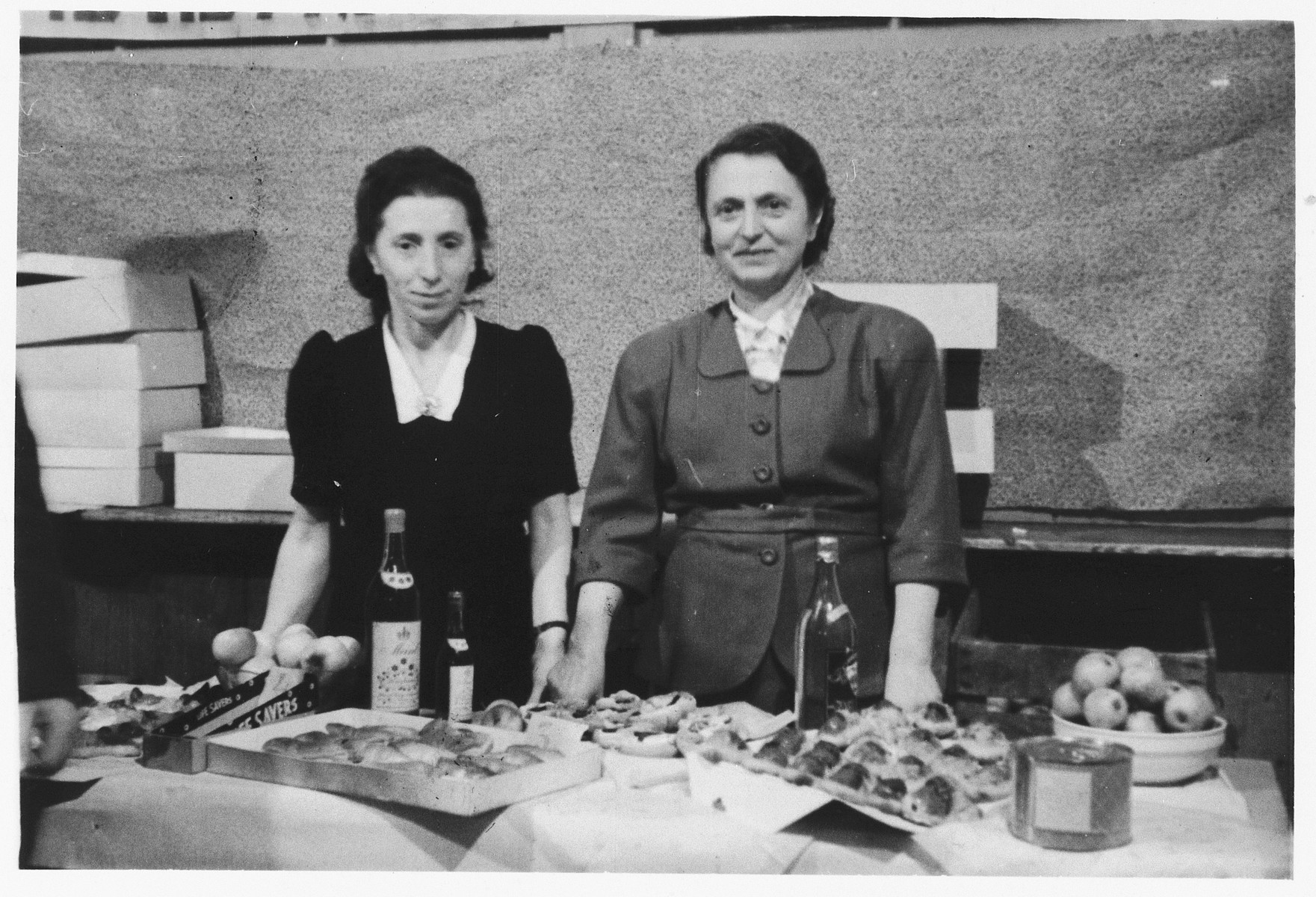 Two women stand behind a table laden with wine and delicacies in the Wels displaced persons' camp.

On the right is Chana Wagner, head of WIZO in Wels.