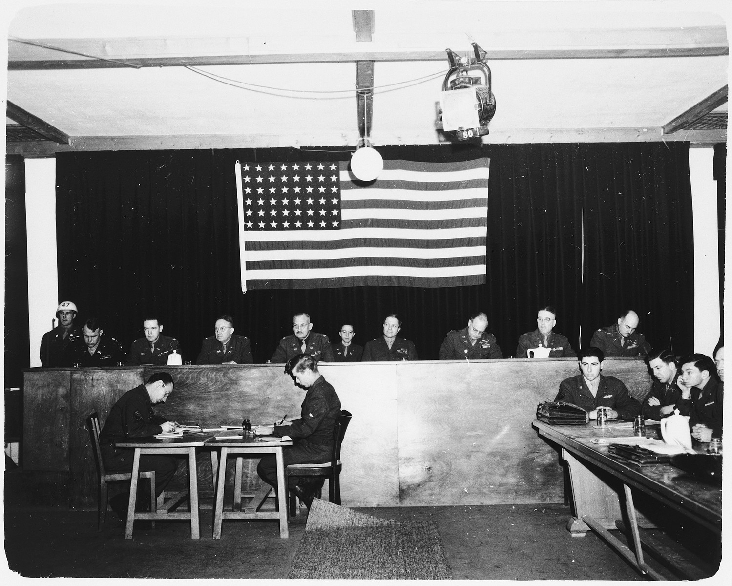 View of the judges bench of the Dachau trial.  A large American flag hangs behind them.

Interpreters for the defense sit at the table to the right of the judges. Among those pictured is Otto Ludwig Stein, third from the left, a 20 year old Sargeant in the US Army. Otto was a Jewish refugee that left Berlin in 1939 and immigrated to the United States. He joined the Army in 1944 and was given the choice of going to Europe or the Pacific, he chose Europe.