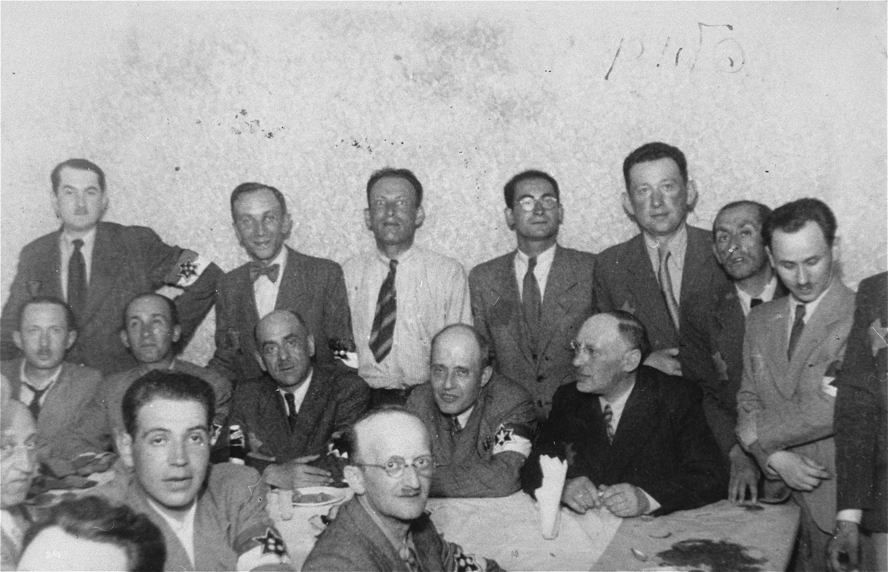 Members of the Lodz ghetto administration, including heads of workshops, police and members of the Sonderkommando, at a social gathering.

Among those pictured are: Leon Fajtlowicz, in charge of the leather workshops (standing fourth from the left), Zygmunt Reingold, director of food supply (standing fifth from the left), Baruch Praszker, chief of special assignments (standing second from the right),  Leon Rozenblatt, chief of the ghettto police (seated on the far side of the table, third from left) and David Warszawski, director of the tailoring workshops (seated on the far side of the table, first from right).