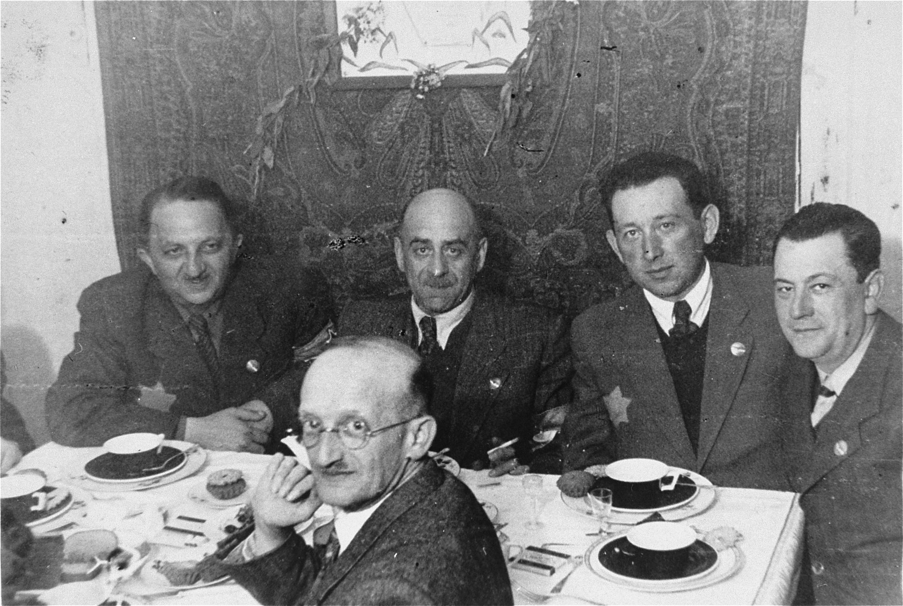 Members of the Lodz ghetto administration are gathered around a table.

Seated on the far side of the table are: Leon Rozenblat, chief of the ghetto police (third from the right), Zygmunt Reingold, head of food supply (second from the right), Jozef Chimowicz, head of the metal workshop and youth employment (far right) and Julek Grosbart (first row) .