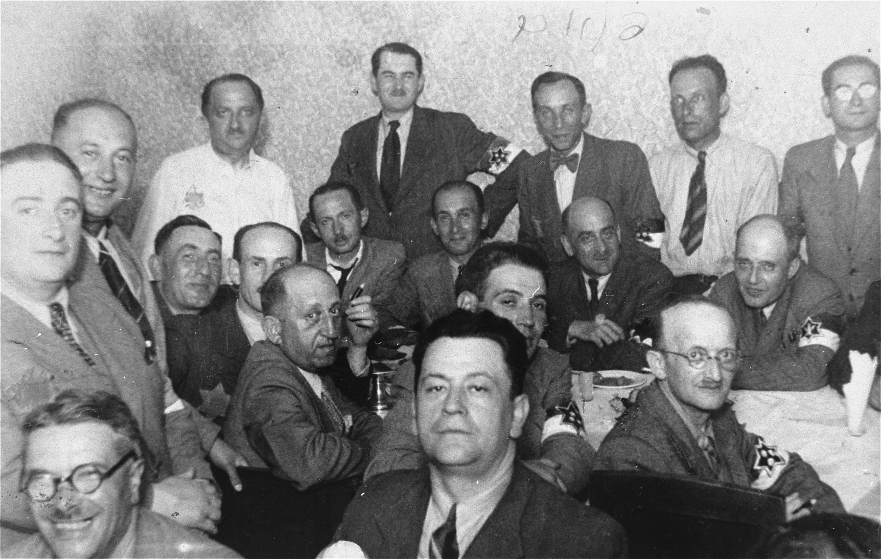 Members of the Lodz ghetto administration, including heads of workshops, police and members of the Sonderkommando, at a social gathering. 

Among those pictured are Abram Josef (Alfred) Chimowicz, head of the housing authority (seated in front, in the center), Leon Rozenblat, chief of the Jewish Police (seated on the far side of the table, second from the right) and Zeligman, head of one of the police precincts (standing in the back row, center).