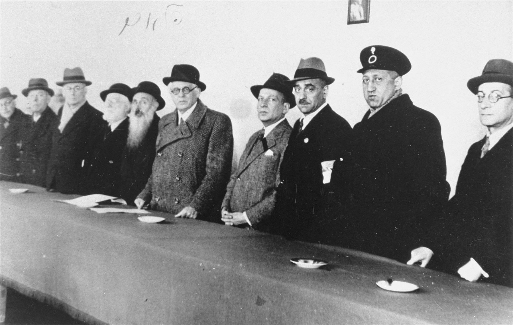 Members of the Lodz ghetto Jewish council stand behind a long table.

Among those pictured are Mordechai Chaim Rumkowski (fifth from the right), Rabbi Yosif Fajner (sixth from the right), Leon Rozenblat (third from the right), Dr. Leon Szykier (second from the right) and Max Szczesliwy.