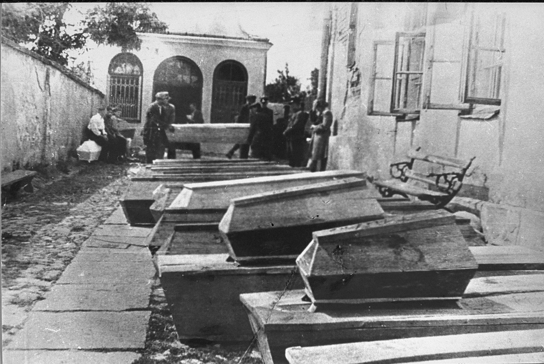 Coffins for the victims of the Kielce pogrom are laid out in a courtyard.  In the background a group of men remove one of the coffins while a young woman holds her head in despair.