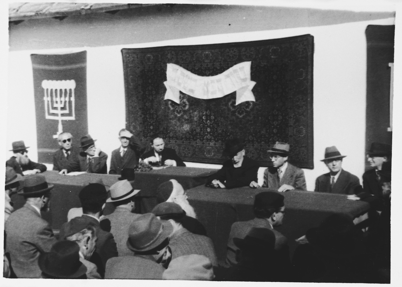 Internees gather for a meeting in the Ferramonti camp to determine work assignments.

This photograph was taken during a visit to the camp by Rabbi Pacifici.

Rabbi Pacifici is seated in the center.  The banner behind him reads "Blessed be your coming in the name of the Lord."