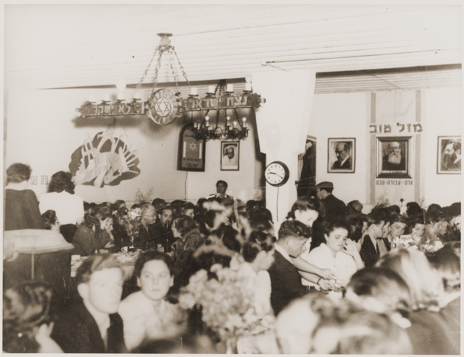 Members of the Kibbutz Nili hachshara (Zionist collective) in Pleikershof, Germany are gathered in the dining hall for a festive meal. 

The room is decorated with portraits of Zionist leaders, including Herzl, Weizmann and A.D. Gordon, and the kibbutz motto.