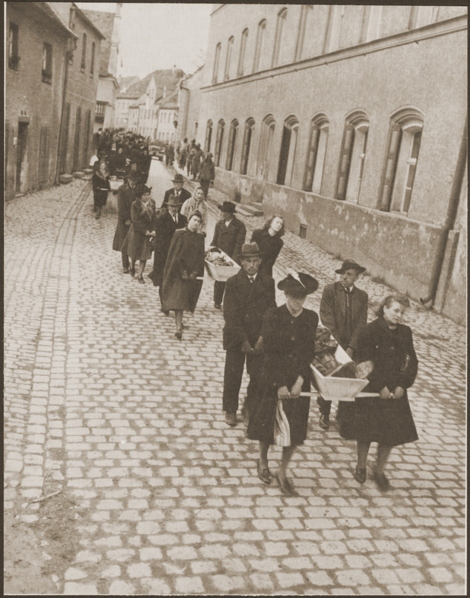 Under the supervision of American soldiers, German civilians from Neunburg vorm Wald carry wooden coffins containing the bodies of Hungarian, Russian and Polish Jews to the town cemetery for proper burial.  

The victims were found in the woods near the town where they were shot by the SS while on a death march from Flossenbuerg.

Original caption reads, "Civilians of the city of Nurnburg at the task of carrying the bodies of the slave laborers over the mile and a half stretch to the burial grounds." [sic]

Original Signal Corps caption reads, "NEUNBURG ATROCITY VICTIMS BURIED.  On orders of Third U.S. Army  officers, German civilians gave a decent burial to 120 Neunburg concentration camp inmates who were murdered by German SS troops in a wooded area one mile from the City of Neunburg before the town was taken by advancing Third Army soldiers.  The victims, Polish and Russian slave laborers, were murdered in several different ways, the most common being by a skull-crushing blow from a blunt instrument.  Some were also shot in the head.  Some of the prisoners were originally from Weimar and several other concentration camps.  Before the atrocity was carried out, German civilians of Neunburg said SS troops ordered citizens of the town into their homes and threatened them with death if they ventured out.  After the Americans took over the area and discovered the crime, they ordered the civilians to carry the corpses from the woodland shooting site into the city for burial in a cemetery.

PNA                                                            EA 64879

THIS PHOTO SHOWS:  German women and men of Neunburg bear the bodies of the victims from the death site to burial in the town cemetery.  U.S. Signal Corps Photo ETO-HQ-45-34602.
SERVICED BY LONDON OWI TO ILST B
CERTIFIED AS PASSED BY SHAEF CENSOR