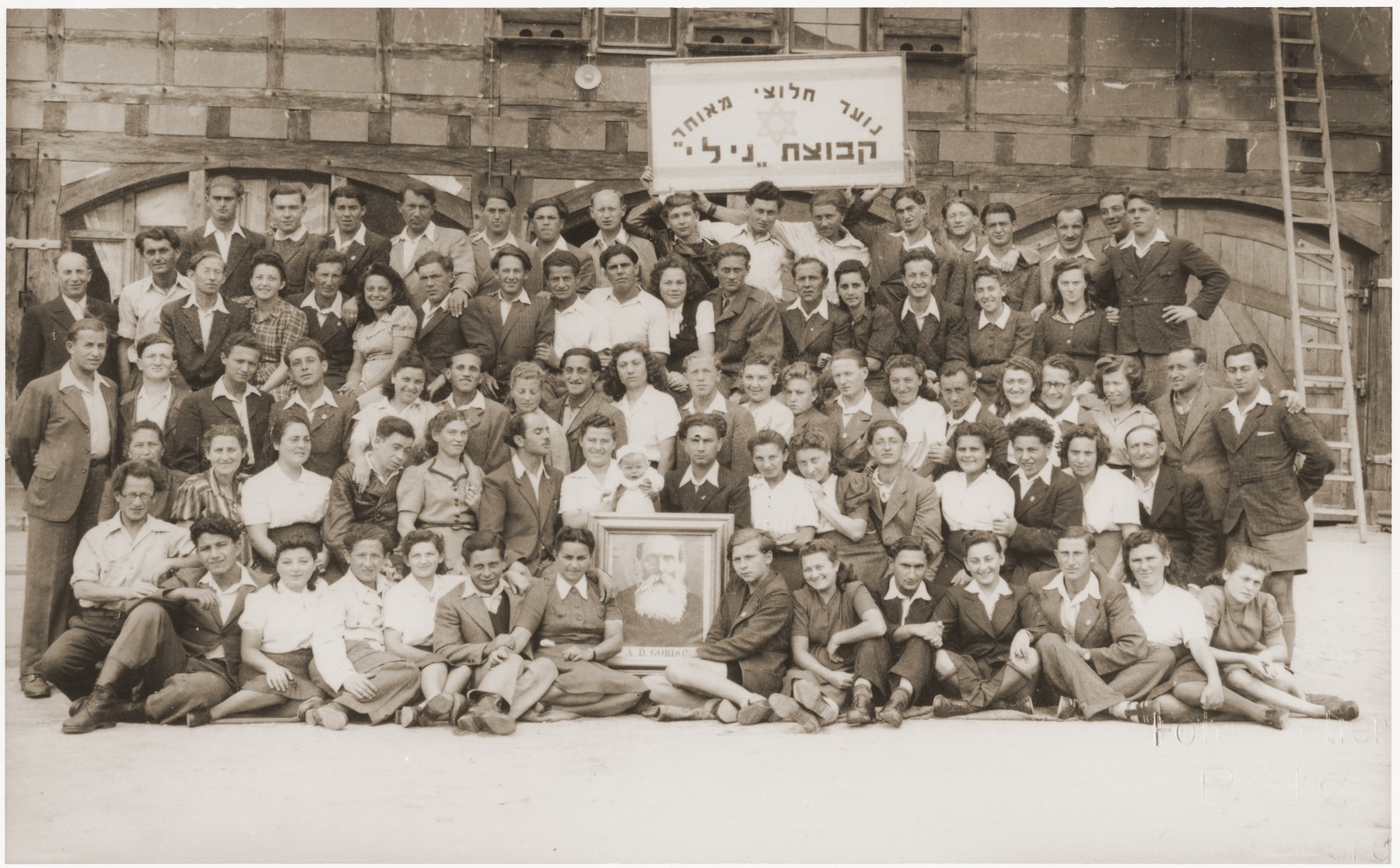 Group portrait of members of the Kibbutz Nili hachshara (Zionist collective) in Pleikershof, Germany

Those in the front row hold a protrait of labor Zionist idealogue A. D. Gordon.  Sitting directly behind the portrait are Noach and Sara (Feldberg) Miedzinski with their infant daughter Nili Ruchana.   Yakov Podolak appears in the second row from the top, eighth 
from the left., and Aryeh Leib Srebrnik is in the second row, far right.
