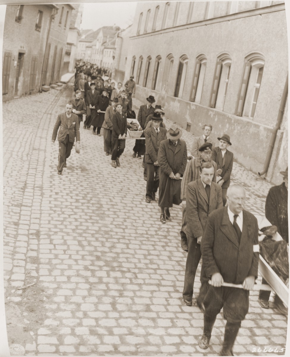 Under the supervision of American soldiers, German civilians carry wooden coffins containing the bodies of Hungarian, Russian and Polish Jews through Neunburg vorm Wald to the town cemetery for proper burial.  

The victims were found in the woods near the town, where they had been shot by the SS while on a death march from Flossenbuerg.