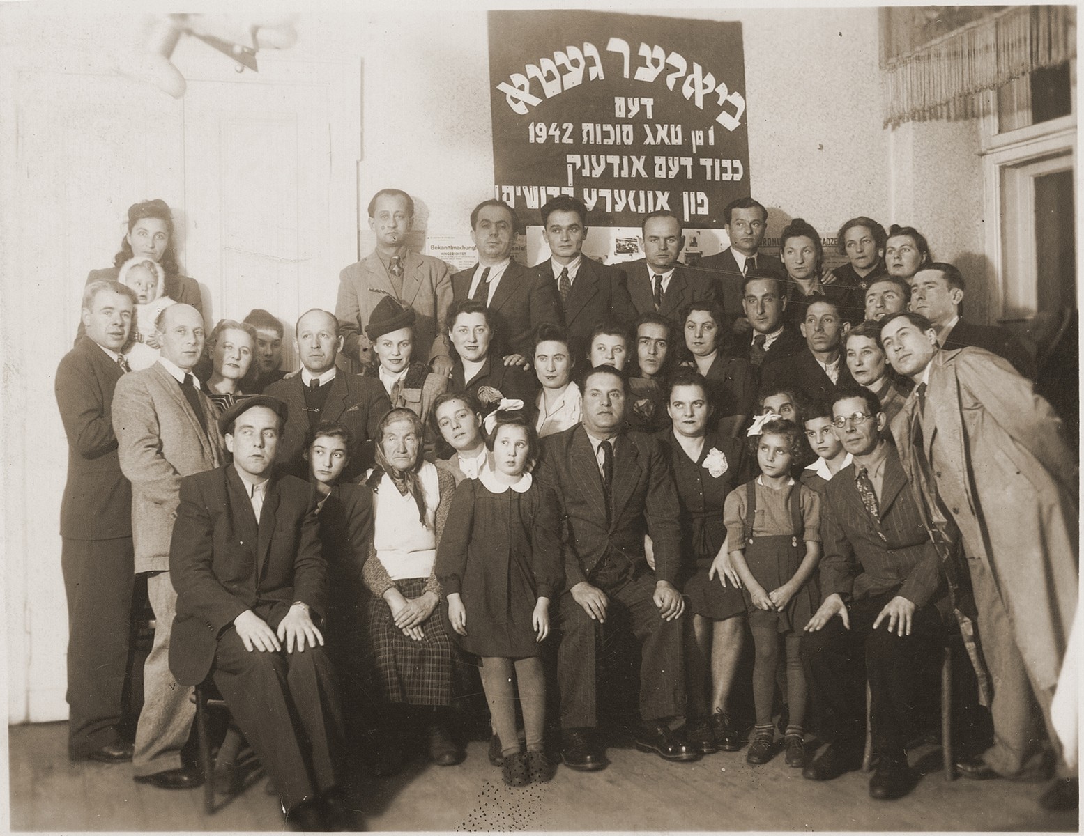 Groups portrait of survivors of the Biala ghetto in front of a Yiddish sign that reads, "Biala Ghetto, the first day of Sukkot 1942.  Honor to the memory of our martyrs."

Gitta Rosenzweig is in the front row, third from right.
