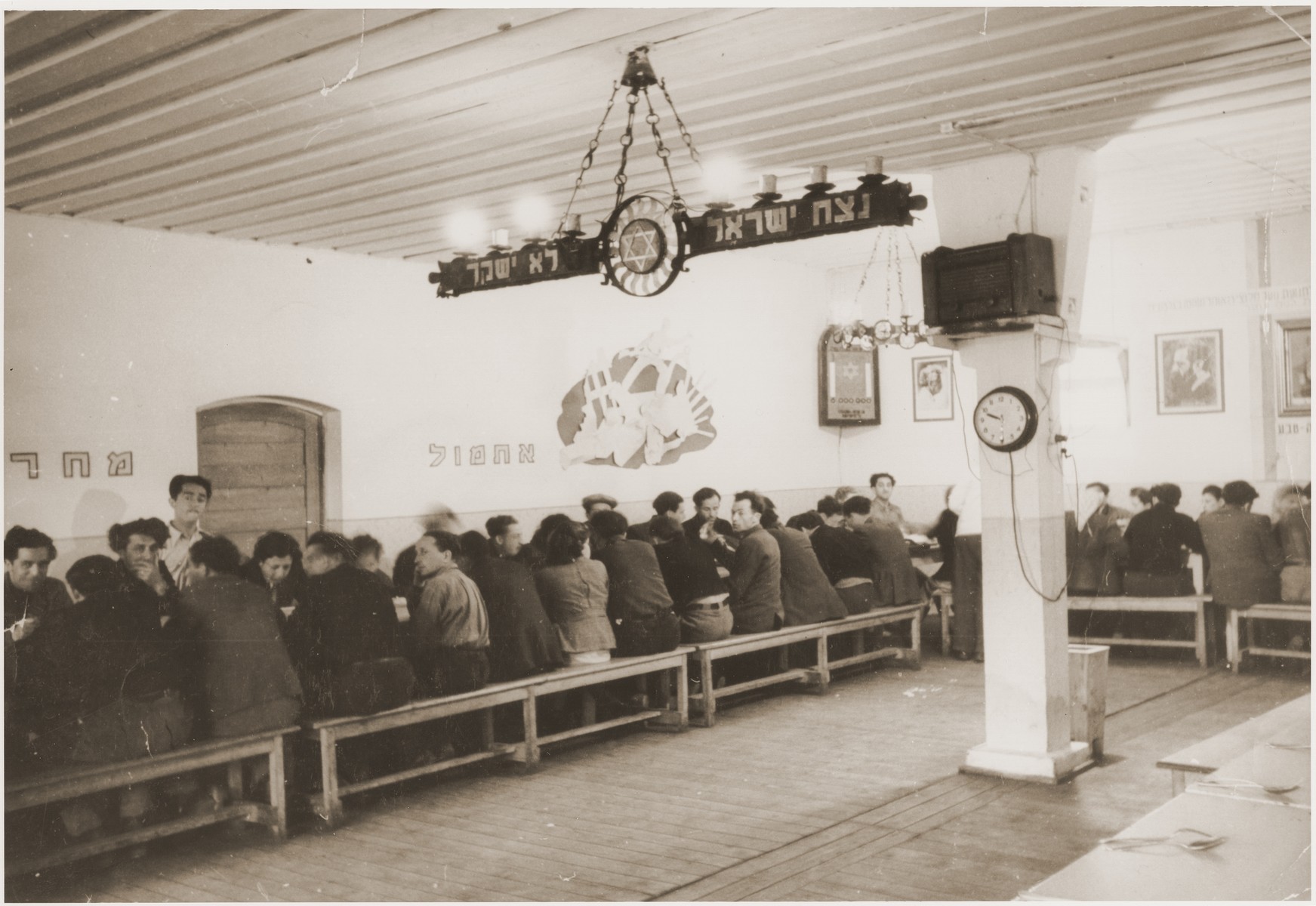 Members of the Kibbutz Nili hachshara (Zionist collective) in Pleikershof, Germany sit around tables in the dining hall.  

The room is decorated with portraits of Zionist leaders, the words "Yesterday" and "Tomorrow" and the motto of the kibbutz.