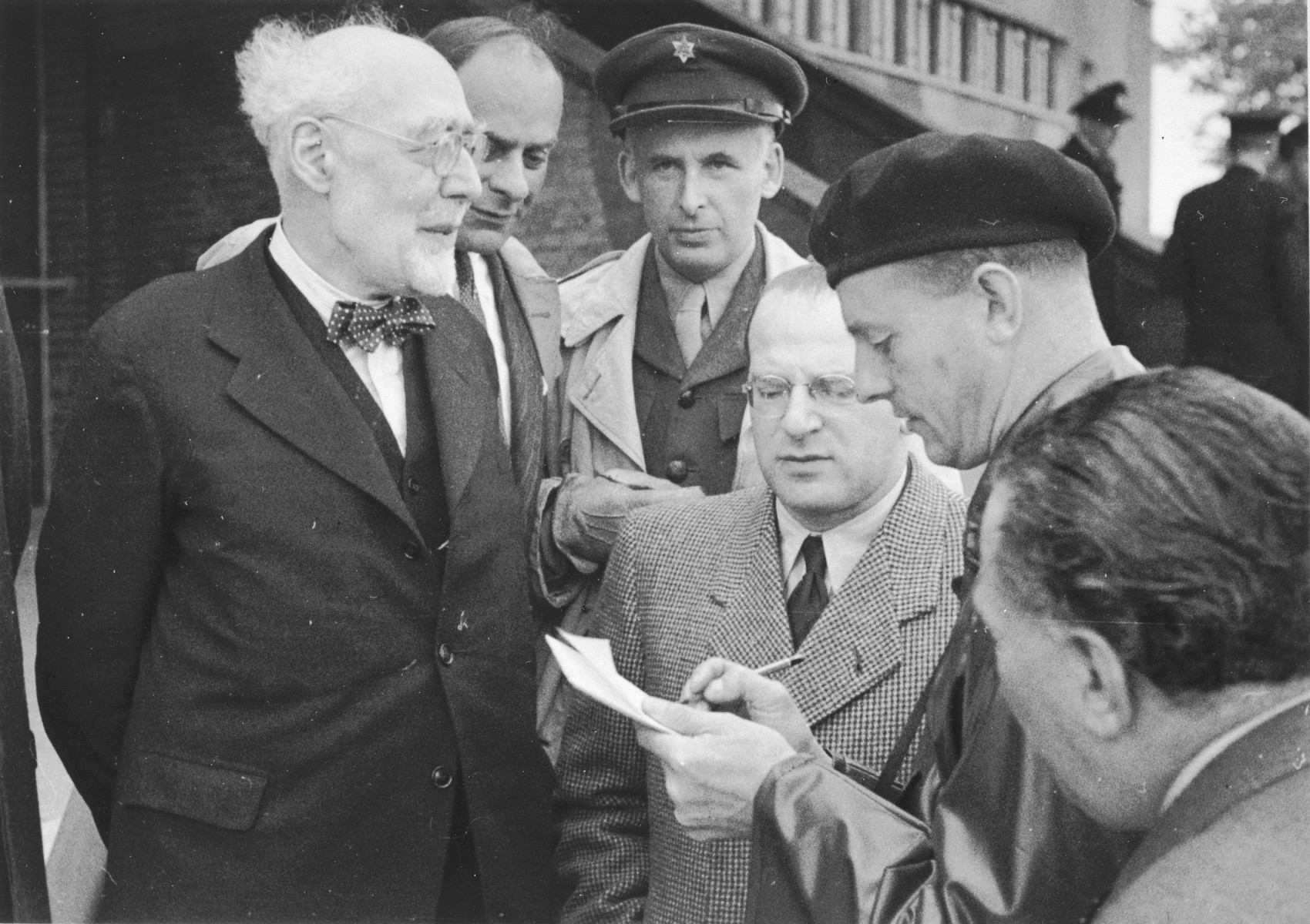 Rabbi Leo Baeck is interviewed by a British soldier upon his arrival in Hamburg at the start of a three week visit to Germany.  

 Also pictured are Norbert Wollheim (wearing glasses in the center) and Heinz Salomon, Leader of Jewish Welfare after the war (behind Leo Baeck).

Baeck was greeted at the airport by Norbert Wollheim, Chairman of the Central Committee of Jews in the British Zone.  Baeck's trip included participation in an evangelical congress in Darmstadt on October 12, as well as numerous appearances before local Jewish communities.