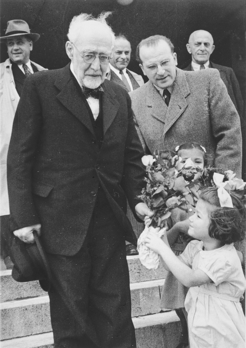 Reha Fabian presents a bouquet of flowers to Rabbi Leo Baeck upon his arrival in Hamburg at the start of a three week visit to Germany.

Reha's sister, Judis, is standing in front of Norbert Wollheim and her father, Hans Erich Fabian, can be seen in the background.

Baeck was greeted at the airport by Norbert Wollheim, Chairman of the Central Committee of Jews in the British Zone.  Baeck's trip included participation in an evangelical congress in Darmstadt on October 12, as well as numerous appearances before local Jewish communities.