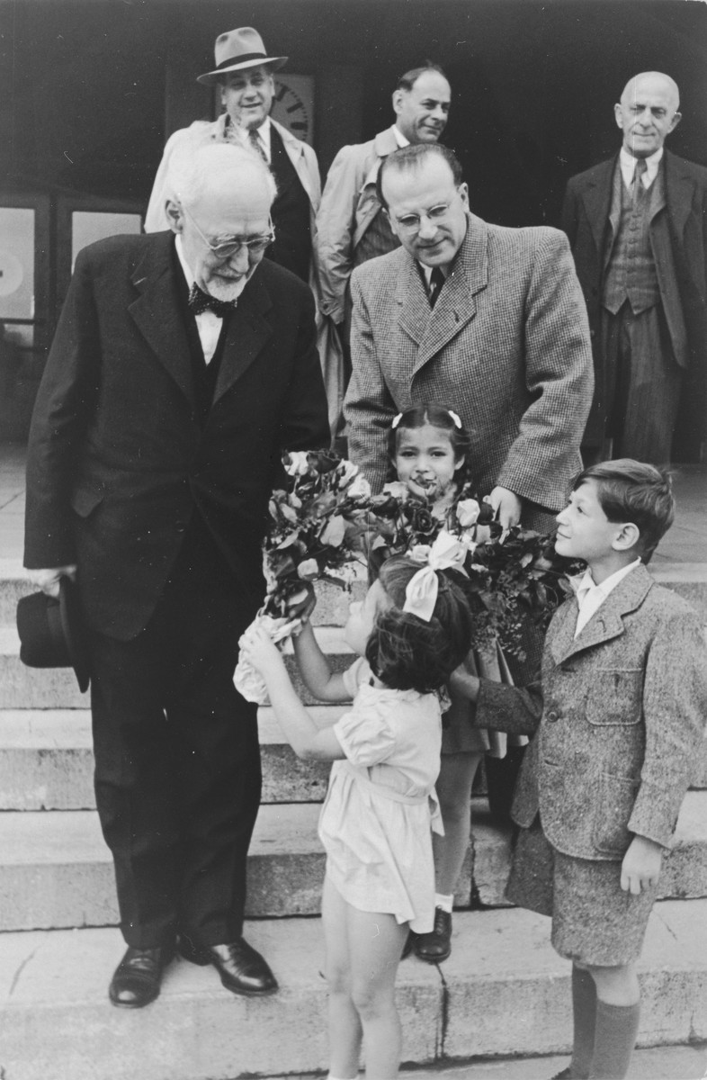 Joel, Reha and Judis Fabian present a bouquet of flowers to Rabbi Leo Baeck upon his arrival in Hamburg at the start of a three week visit to Germany.

The three children are survivors of Theresienstadt.  Their father Hans Erich Fabian is standing in the background wearing a hat.

Baeck was greeted at the airport by Norbert Wollheim, Chairman of the Central Committee of Jews in the British Zone.  Baeck's trip included participation in an evangelical congress in Darmstadt on October 12, as well as numerous appearances before local Jewish communities.