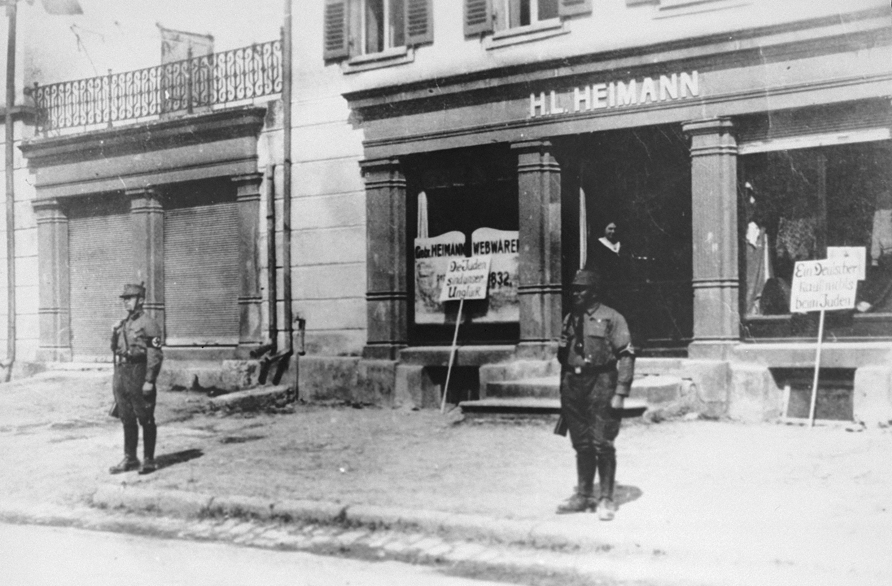 Two Nazi stormtroopers stand guard in front of the H. L. Heimann store in Bopfingen, to prevent would-be shoppers from violating the Nazi boycott of Jewish-owned businesses.  Two signs have been placed in front of the store.  The sign on the left reads: "The Jews are our Misfortune."  The sign on the right reads:  "A German buys nothing from Jews."

The business belonged to the donor's father.  Pictured at the entrance to the store is Sali Heimann, who was later killed in a concentration camp in Riga.