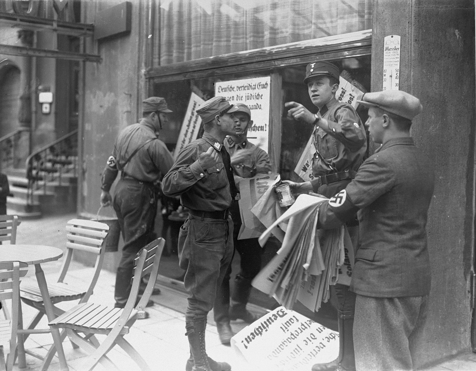 An SA member instructs others where to post anti-Jewish boycott signs on a commercial street in Germany.  

A German civilian wearing a Nazi armband holds a sheaf of anti-Jewish boycott signs, while SA members paste them on a Jewish-owned business.  Most of the signs read, "Germans defend yourselves against Jewish atrocity propaganda/Buy only at German stores."