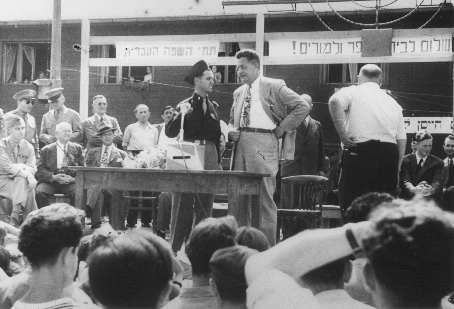U.S. Army chaplain Rabbi Mayer Abramowitz converses with an official at an outdoor forum in the Schlachtensee displaced persons camp.

The forum was held for the benefit of a visiting international commission.  Above the podium is a sign in Hebrew that reads, "Long live the Hebrew language."