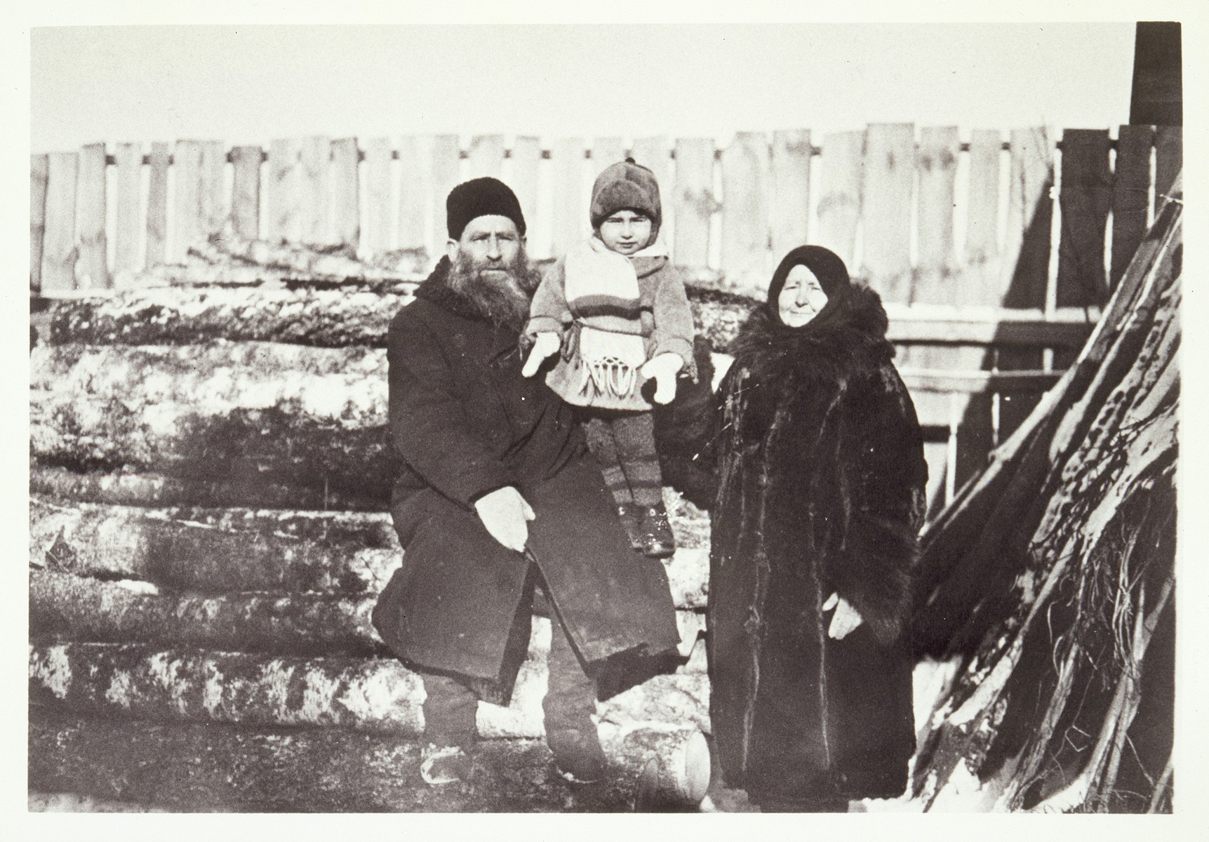 Reb Dovid and Leah Moszczenik pose by a wood pile with their grandson Yudaleh Kabacznik in the family's backyard. 

Yudaleh was killed by the Polish Home Army in 1944. Leah was killed by the Germans during the September 1941 mass shooting action in Eisiskes.  Reb Dovid died a natural death.