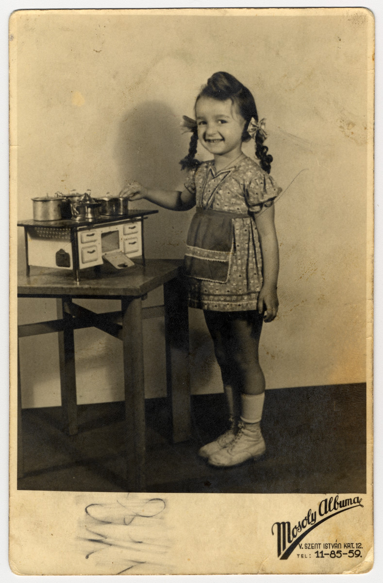 Sutdio portrait of a Hungarian Jewish girl standing next to a toy kitchen and pretending to cook.

Pictured is Zsuzsa Rubin.  This photograph was sent to her uncle Izso Stern in a Hungarian labor battalion.  One month after this photograph was taken, Germany seized control of Hungary and began implementing the Final Solution there.