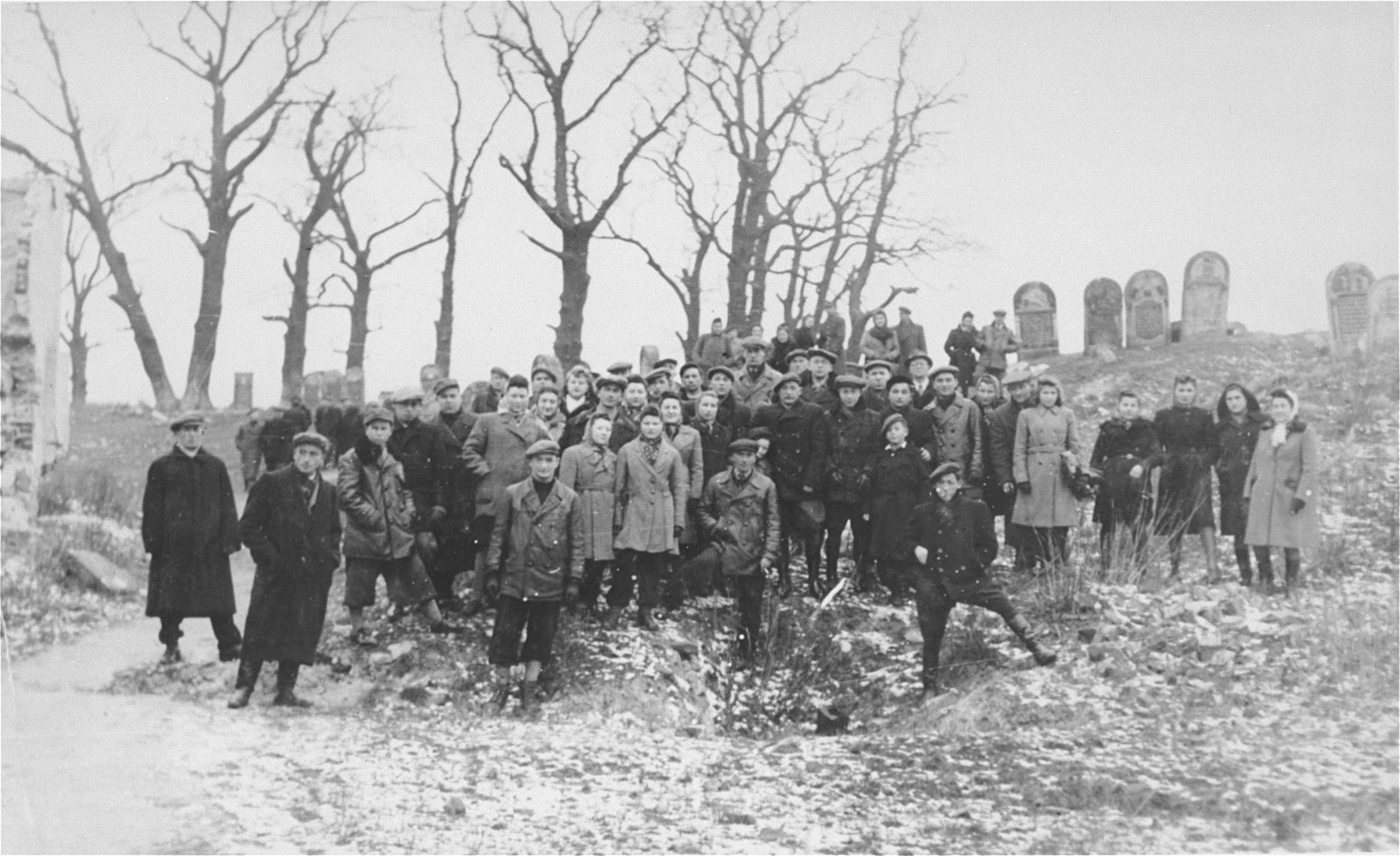 Group portrait of Jews from Ostrowiec standing on the site of a mass grave for 2000 Jews shot during the October 1942 action.  Behind them is a portion of the vandalized Jewish cemetery.  

Among those pictured is Berek Blumensztok.  He is standing in the center. Berek  returned to Ostrowiec after the war with the hope of reuniting with his family.  However, all had perished, including his mother, father and 8 siblings. 

Also pictured is Dora Weinberg Berman (third woman from the right).