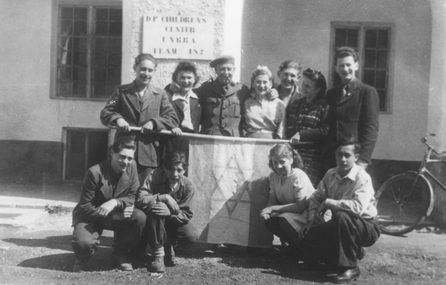 A group of youth pose by a Zionist flag in the International DP Children's Center Kloster Indersdorf 

Pictured kneeling on the far left is Erwin Farkas; kneeling on the far right is Moniek (Manny) Drukier; Iwan Kicz (Irving Klein) is in the back row, third from the right; and standing in the center is Mr. Parker.