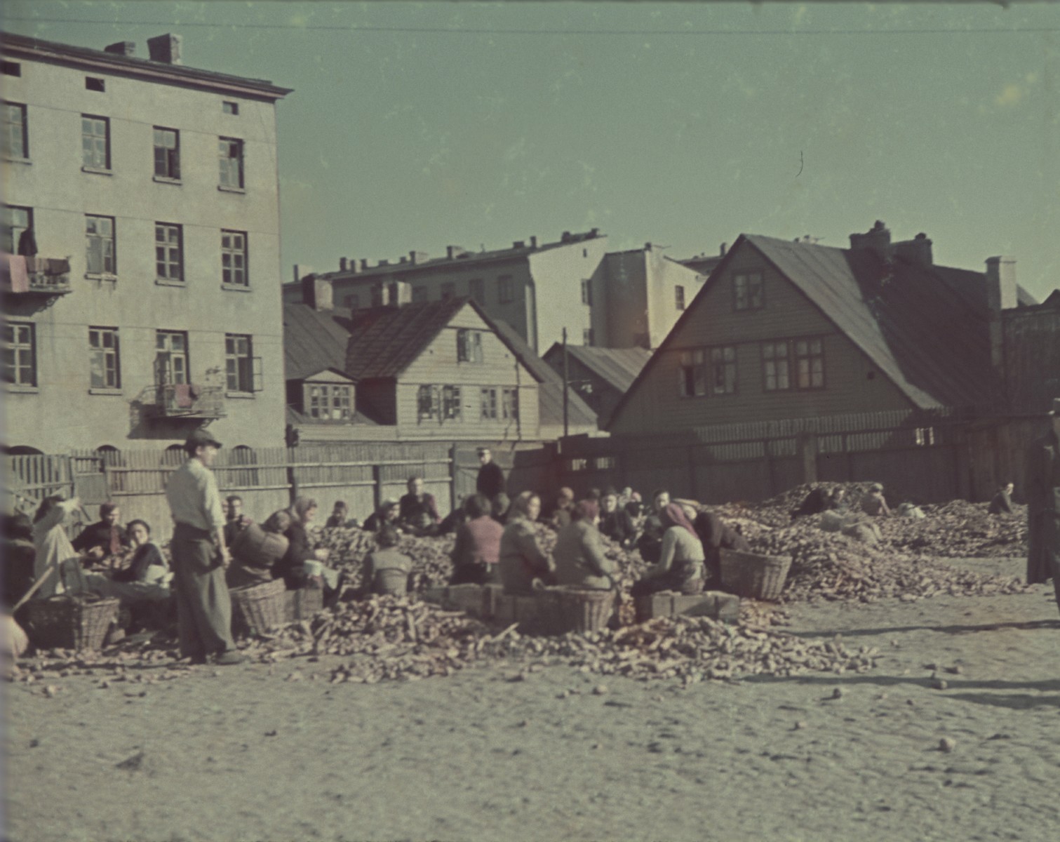 Women sit on crates in a large open square and sort out vegetables.

Original German caption: "Lagerplatz" (camp square) #143.