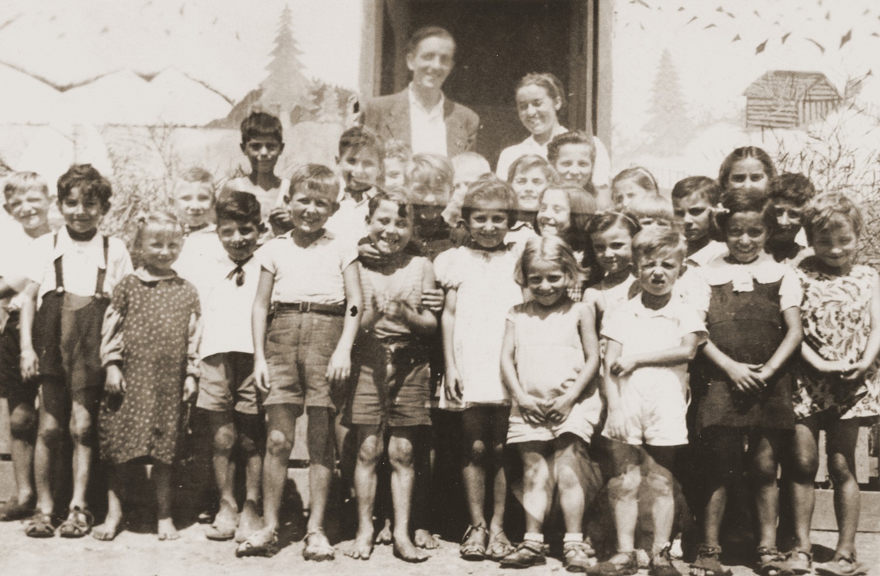 Friedel Reiter and August Bohny pose with a group of children in front of the Secours Suisse barracks in the Rivesaltes internment camp.  

The building is decorated with a mural of the Swiss Alps.
