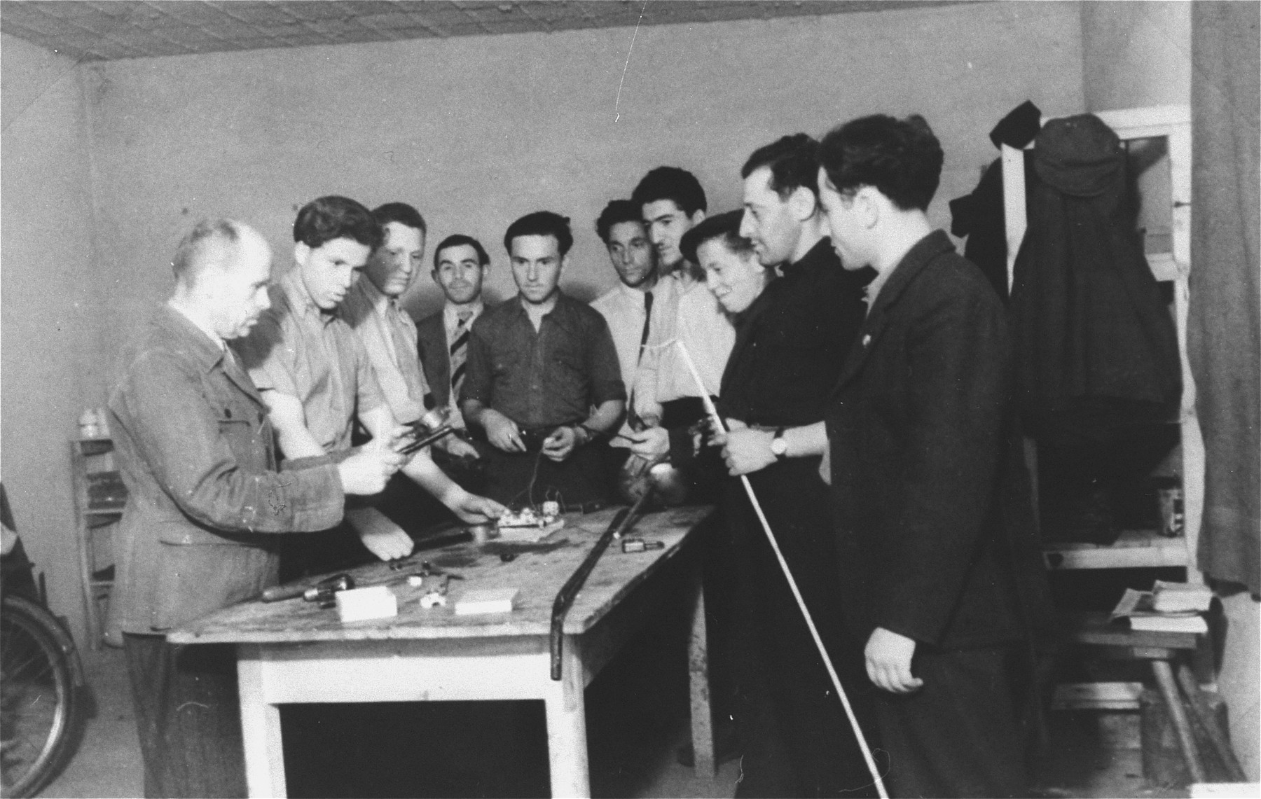 Men gather around a table for a tools demonstration in an ORT sponsored vocational school in the Zeilsheim displaced persons' camp.

Pictured among the men in the background is Jana Wisgardisky.