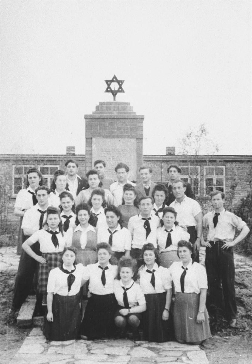 Group portrait of a Zionist youth organization in Zeilsheim standing in front of a memorial to victims of the Holocaust.

Pictured among the group is Sheyne-Bela Berk (Propis).