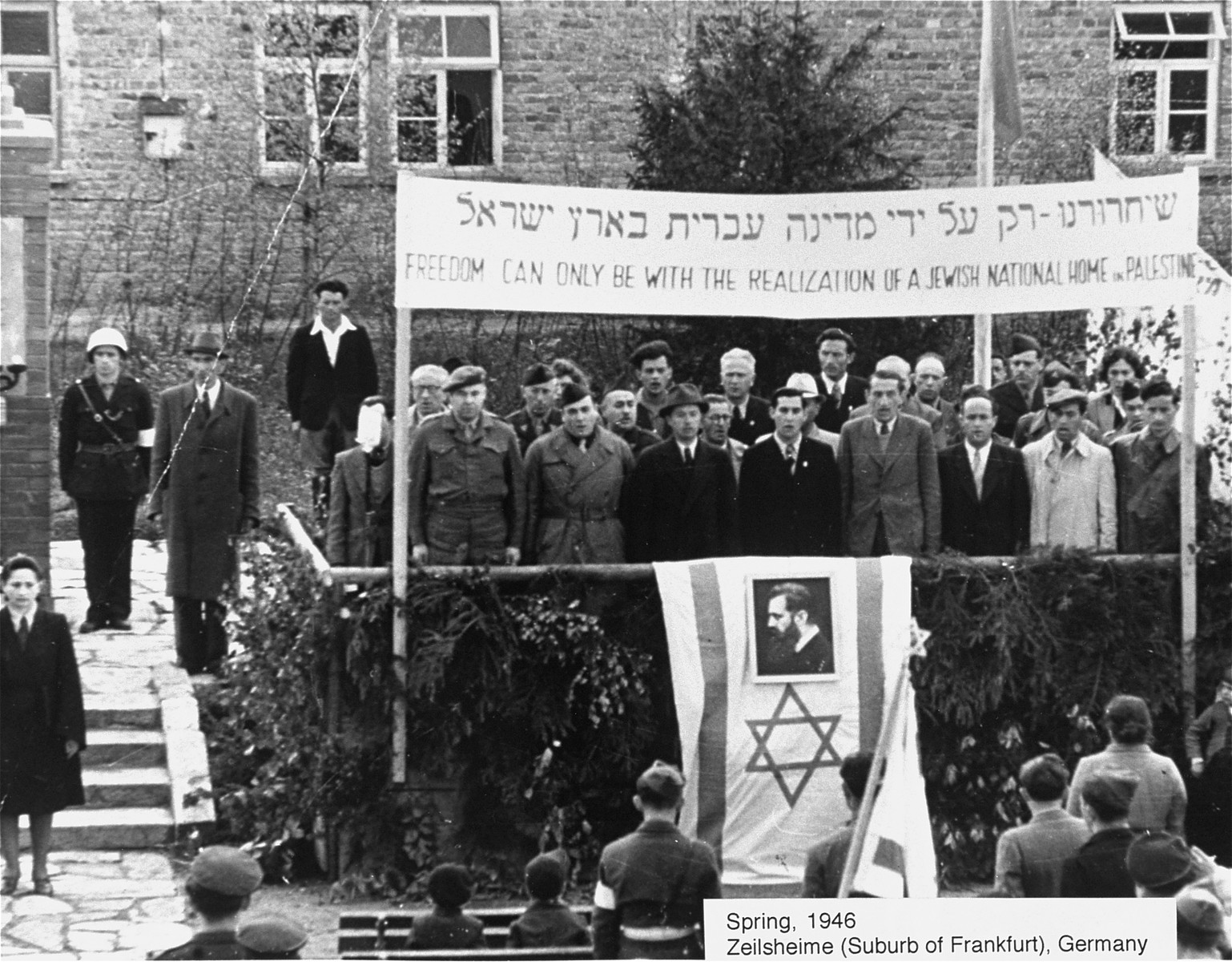 Camp administrators and Zionist leaders, standing beneath a large banner, sing  the Jewish national anthem (Hatikva) at a Zionist rally at the Zeilsheim displaced persons camp.