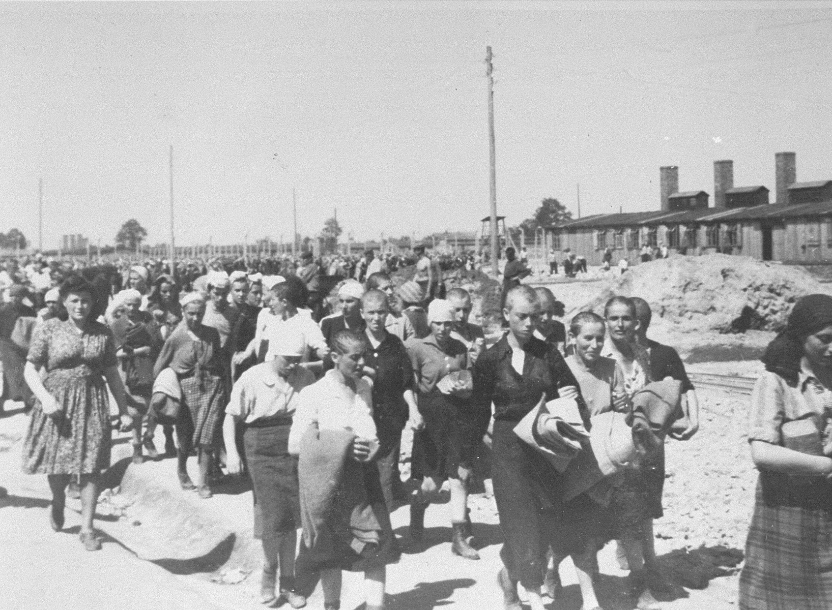 Jewish women from Subcarpathian Rus who have been selected for forced labor at Auschwitz-Birkenau, march toward their barracks after disinfection and headshaving.  

The woman on the left with a full head of hair is the Blockaelteste.  The building on the right is the kitchen of the women's camp.  Pictured in the back is Hildegard Taussig.
