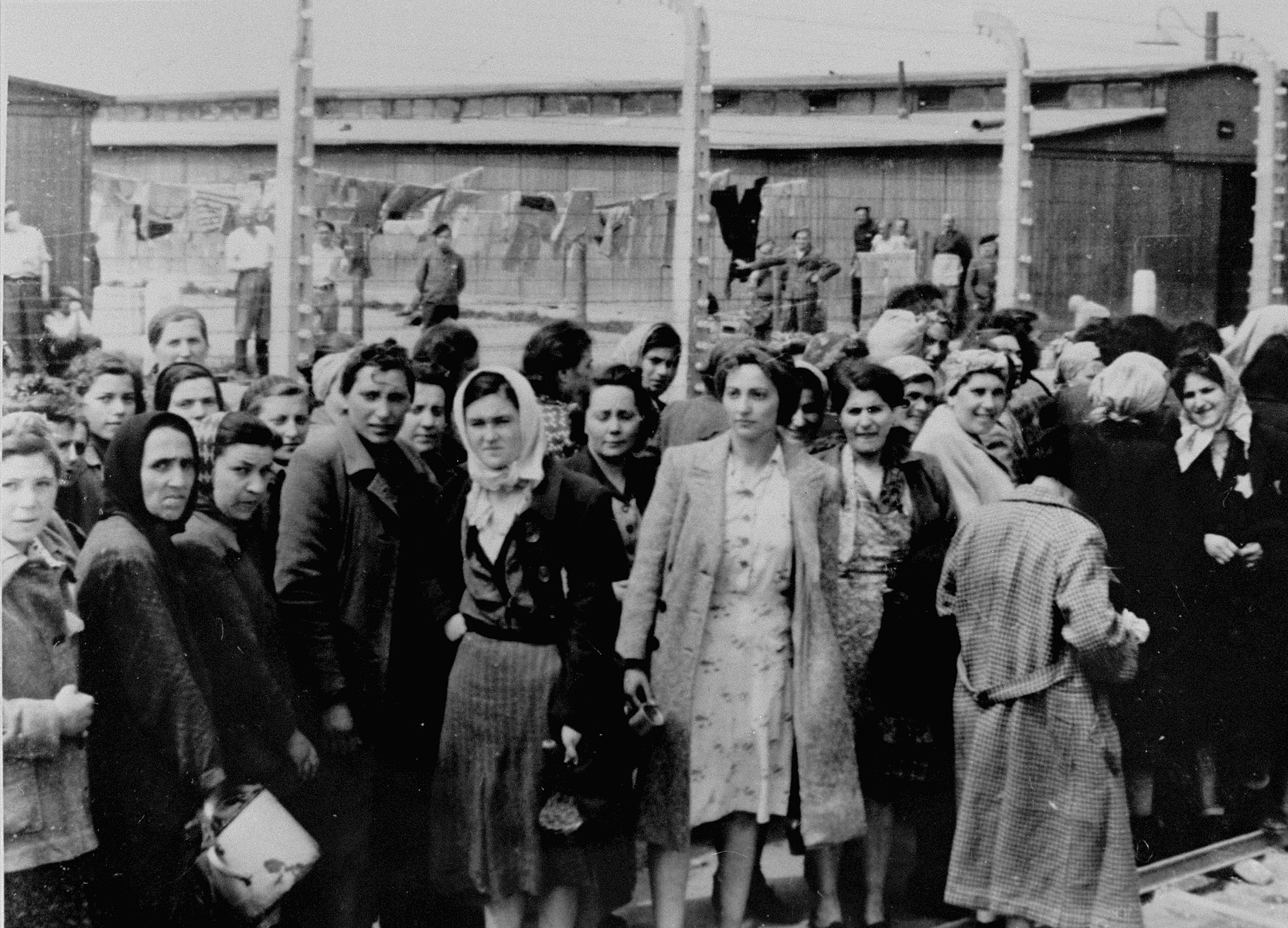 Jewish women from Subcarpathian Rus who have been selected for forced labor at Auschwitz-Birkenau, wait to be taken to another section of the camp.
 
Behind the fence is block BIId where 20,000 men were imprisoned. 

Among those pictured are Gizi Krousz Kauf of Szeged, Rozsi Egri Swartz, Zseni Stern of Bodrogkeresztur, Ester Erzsebet Egri Deutsch (Rozsi's niece) of Bodrogkeresztur, Freda Egri Salomon (Ester's sister), Aranka Stern (Zseni's niece) and Etu Szumet of Bodrogkeresztur.