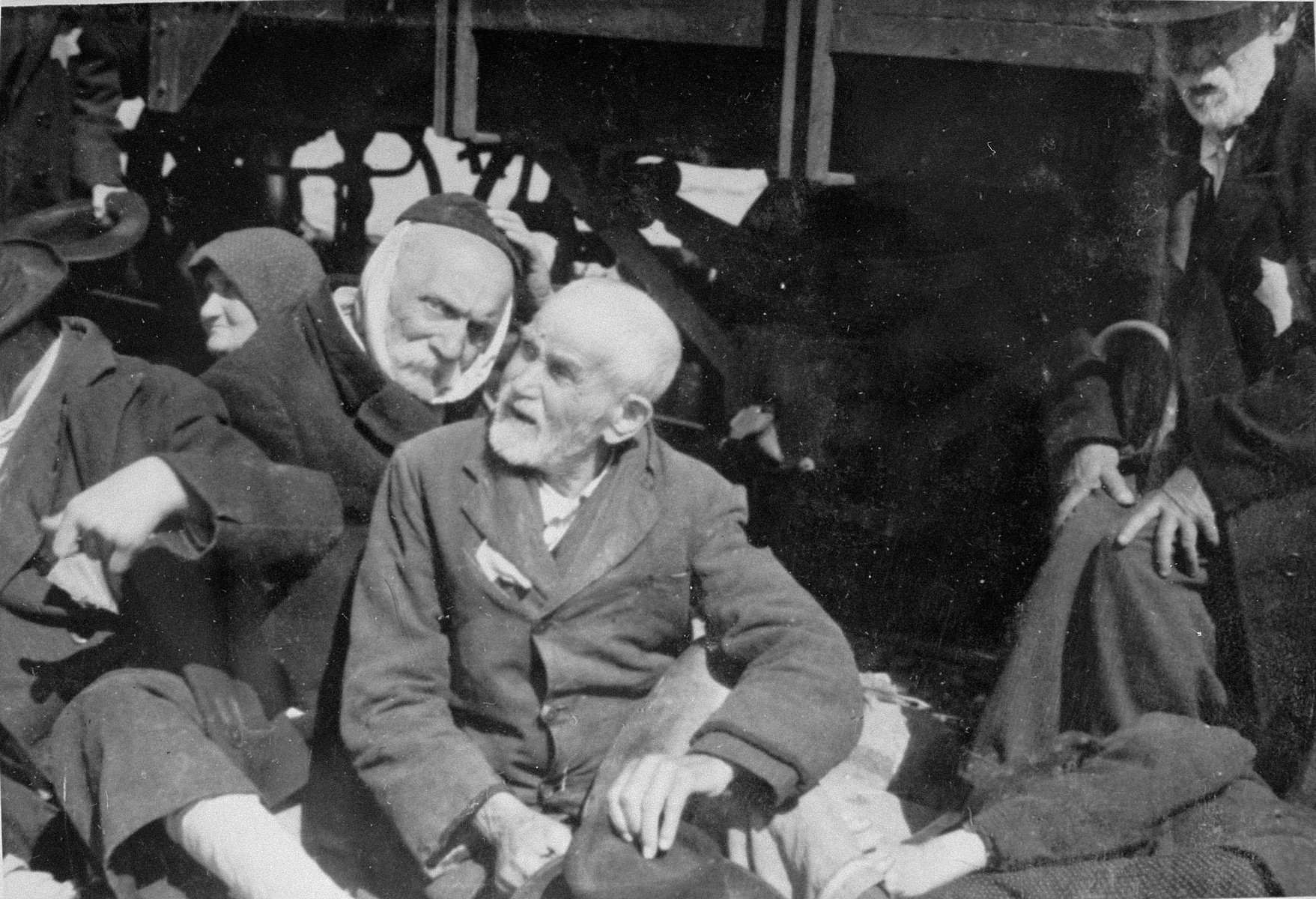 Elderly Jewish men from Subcarpathian Rus sit next to a railcar in Auschwitz-Birkenau prior to being sent to the gas chambers. 

Seated on the left is Henryk Naftoli Herz Adler.  He is wearing a handkerchief to cover his face because his beard was forcibly cut off.  Seated at the right is Samuel (Sandor) Ben Yehuda Kohn. The man on the far right is Lili Jacob's paternal grandfather.
